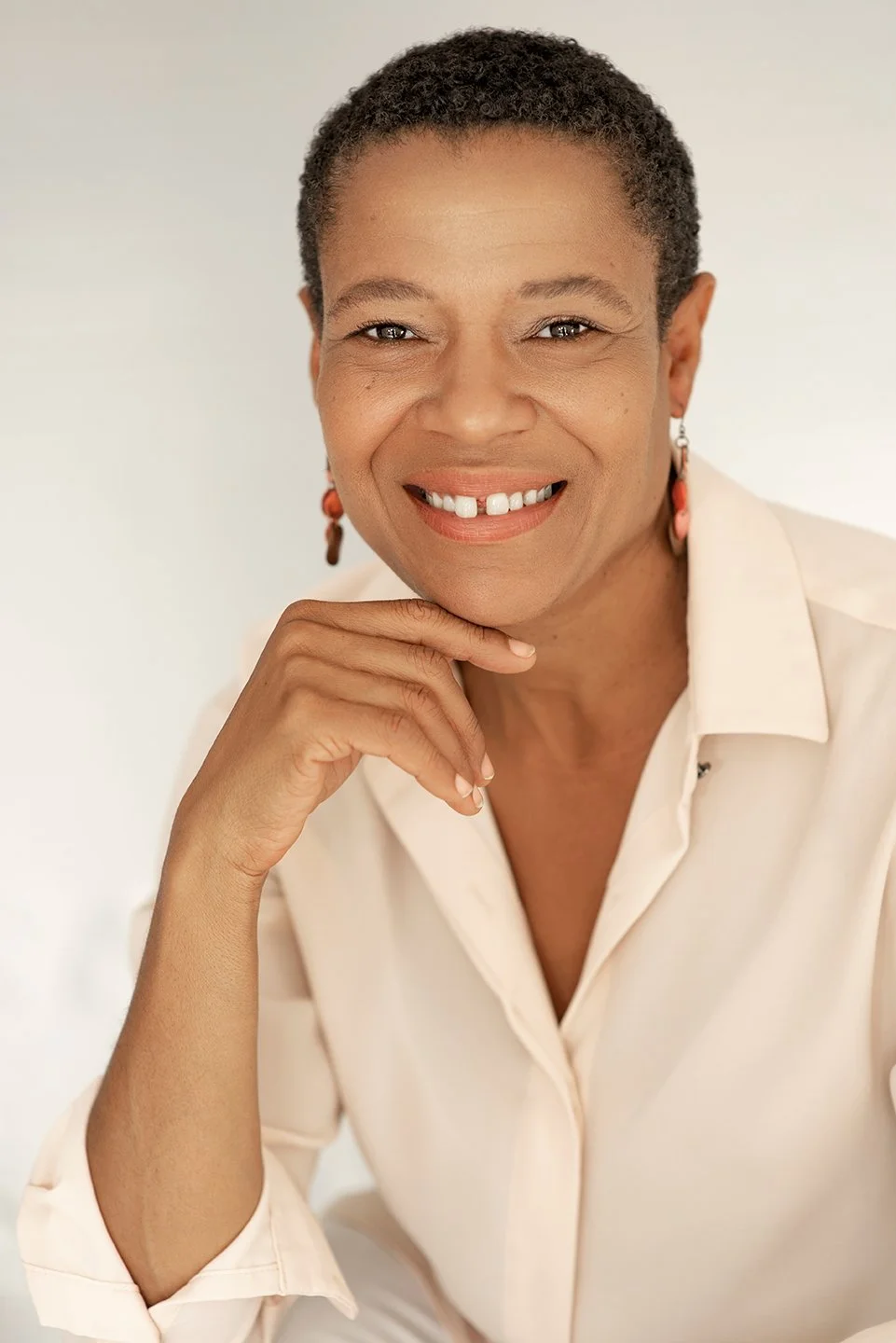 A smiling middle-aged African American woman with short curly hair, wearing earrings and a cream-colored collared shirt, posing against a plain light background.