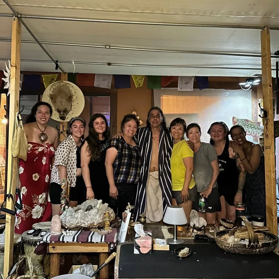 Group of people smiling and posing together inside a decorated room with native and cultural elements, including a large straw hat and colorful banners.