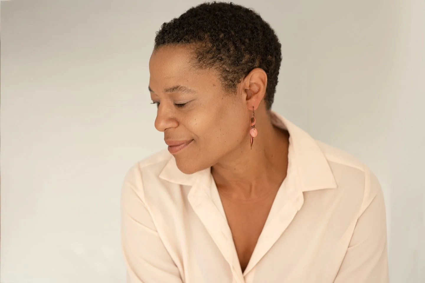 A woman with short curly hair and earrings, wearing a light-colored shirt, looking down with a gentle smile.