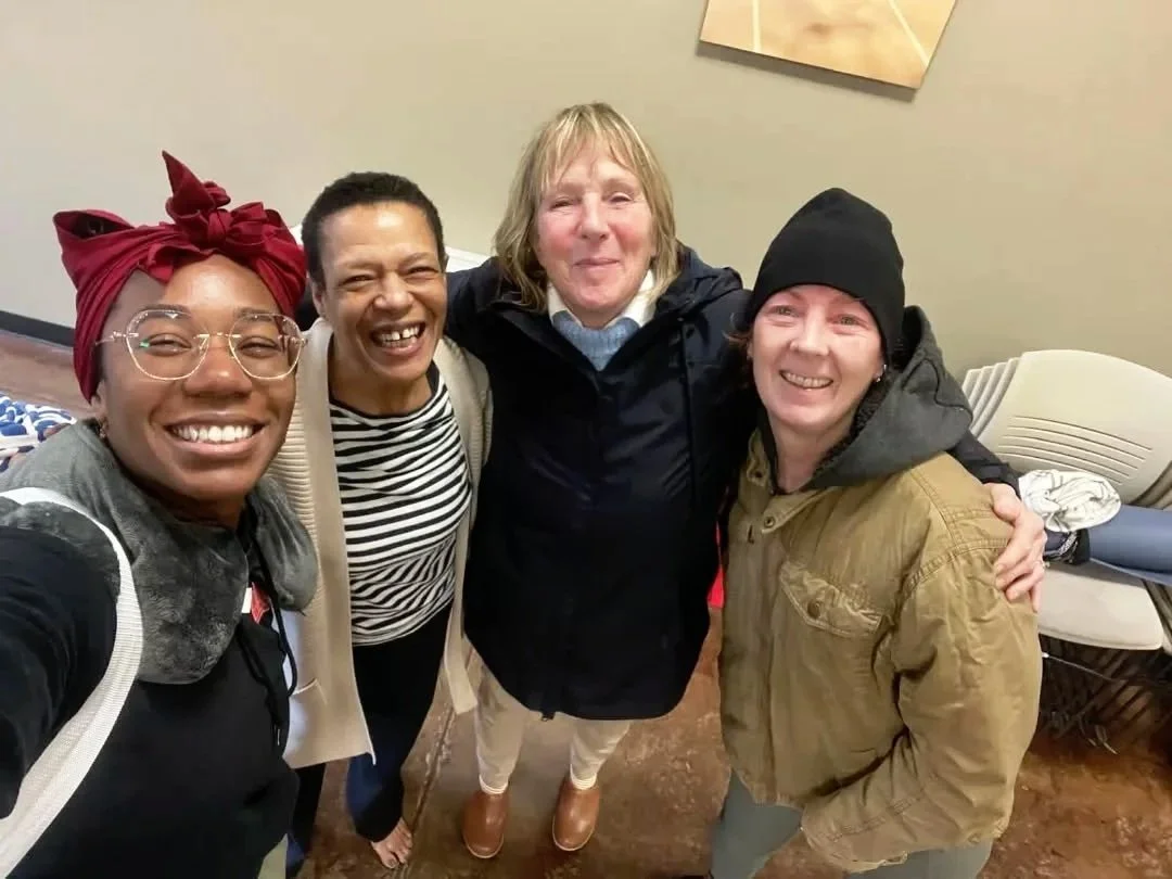 Four women standing closely together indoors, smiling for a selfie, with chairs and a wall in the background.
