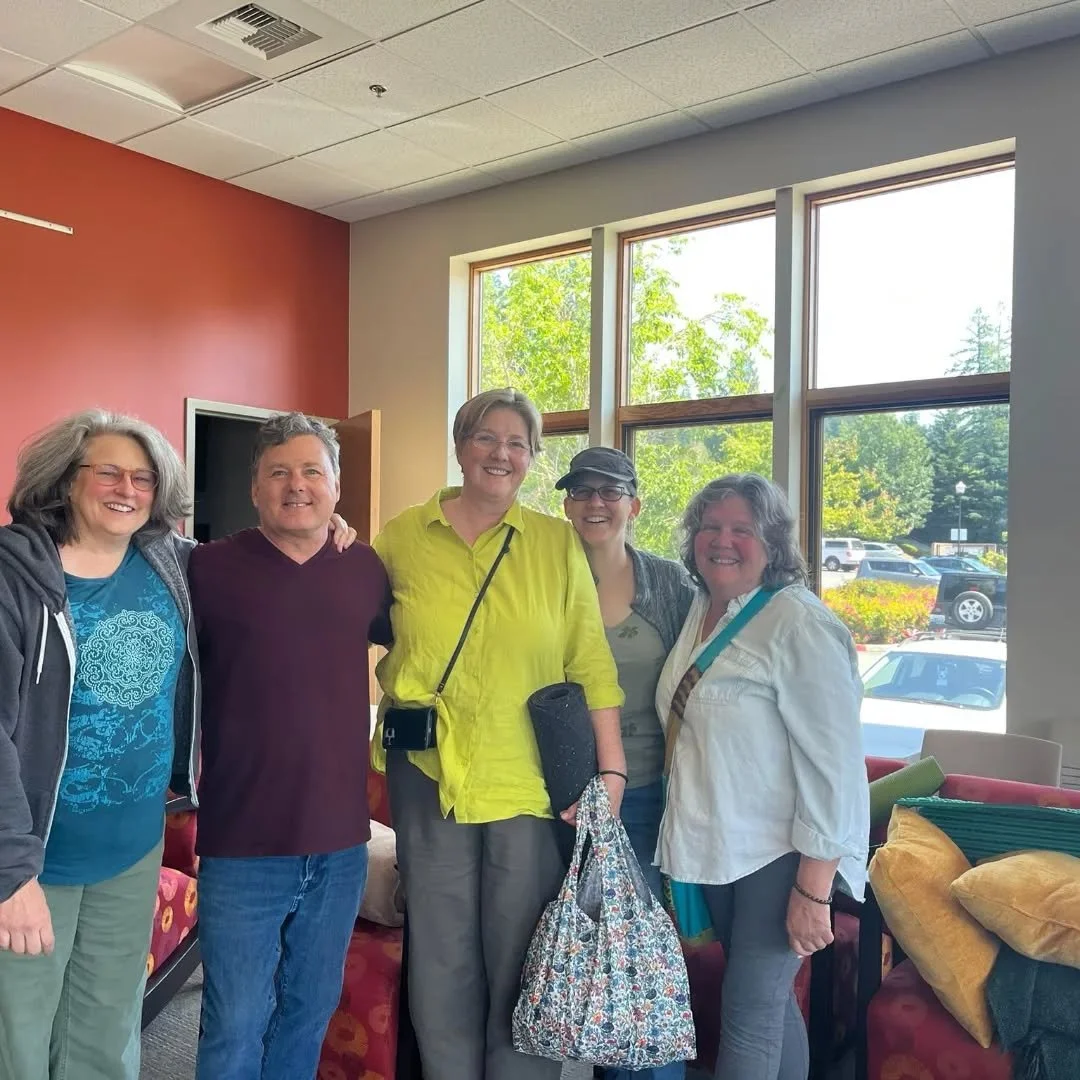 Five women standing indoors near large windows, smiling for a group photo.