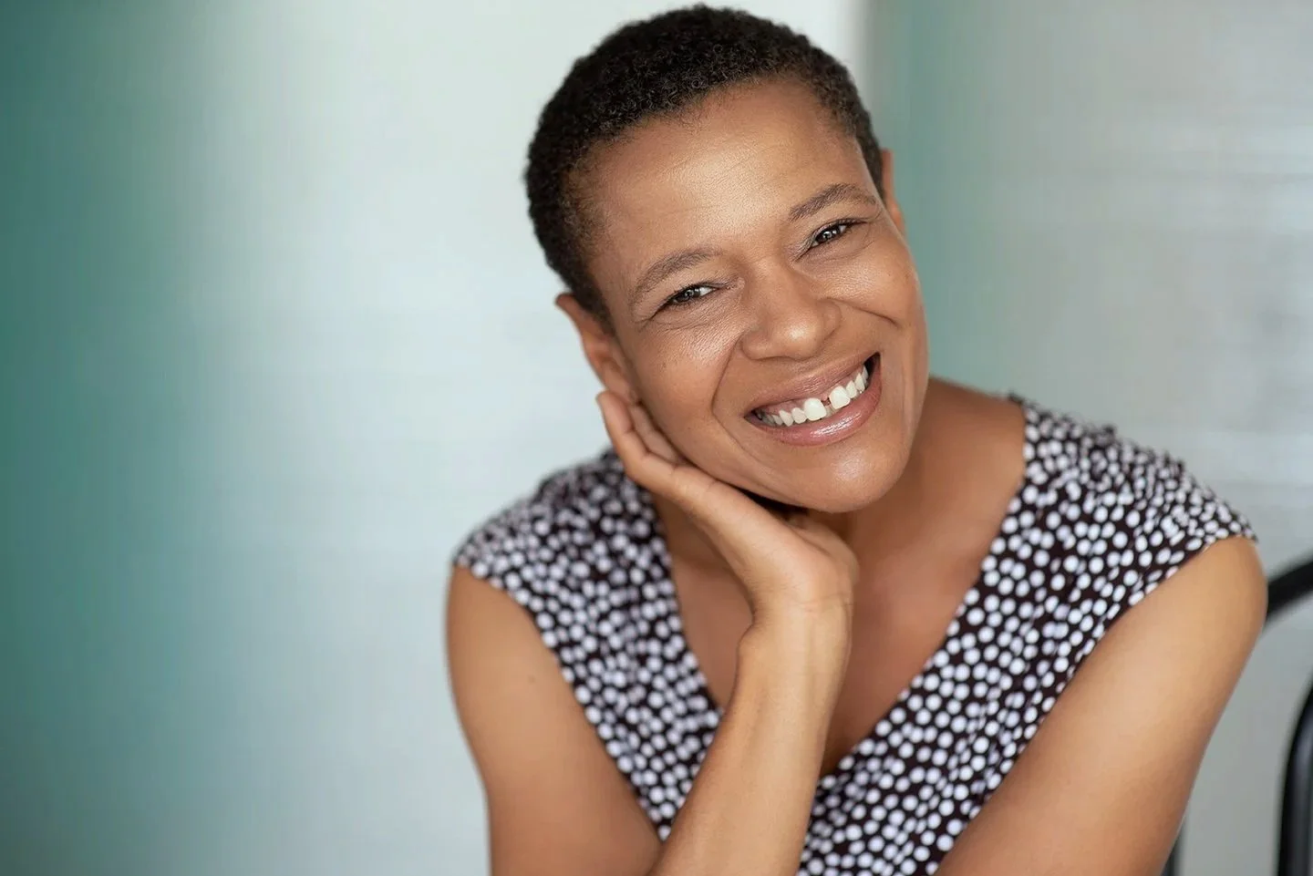 A woman with short curly hair wearing a black and white polka dot top, smiling, resting her face on her hand.