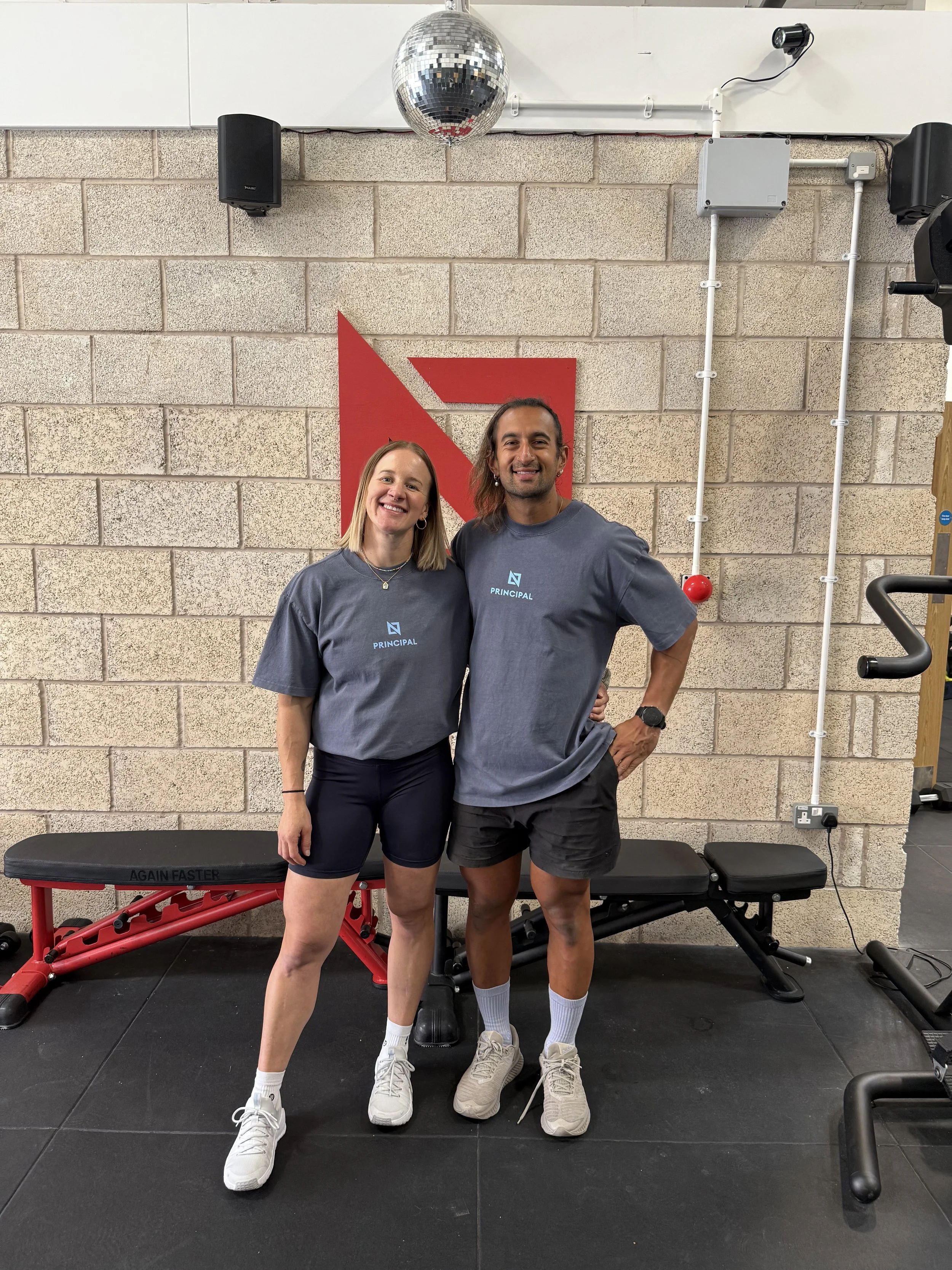 Two smiling people standing in a gym, wearing matching gray shirts with a logo and the word 'Principal'. They are in front of industrial-looking walls, with gym equipment including a massage table, weight bench, and a vertical tension-based exercise machine nearby, and a disco ball hanging from the ceiling.