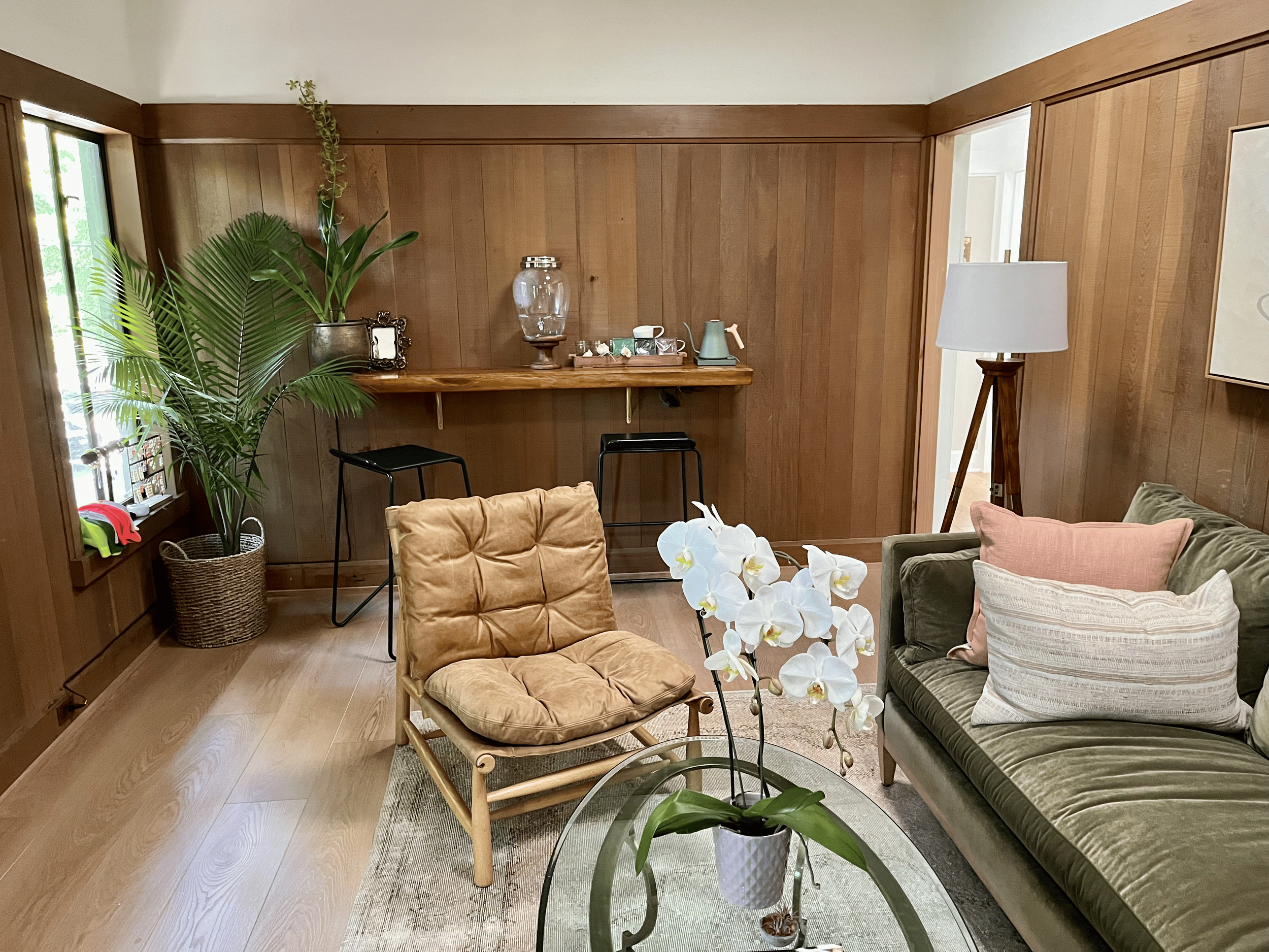 Living room with wooden walls, large plant near window, beige and green seating, white orchids in a gray pot, standing lamp, and a wooden table with decorative items including a water dispenser, teapots, and cups.