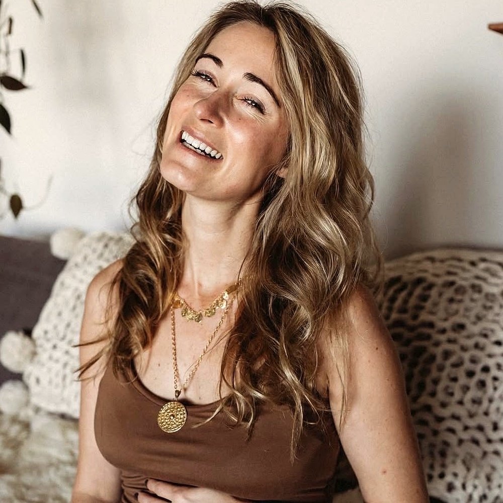 A woman with curly blonde hair smiling and wearing layered gold necklaces and a sleeveless brown top, sitting on a sofa with leopard print and peacock feather pillows.