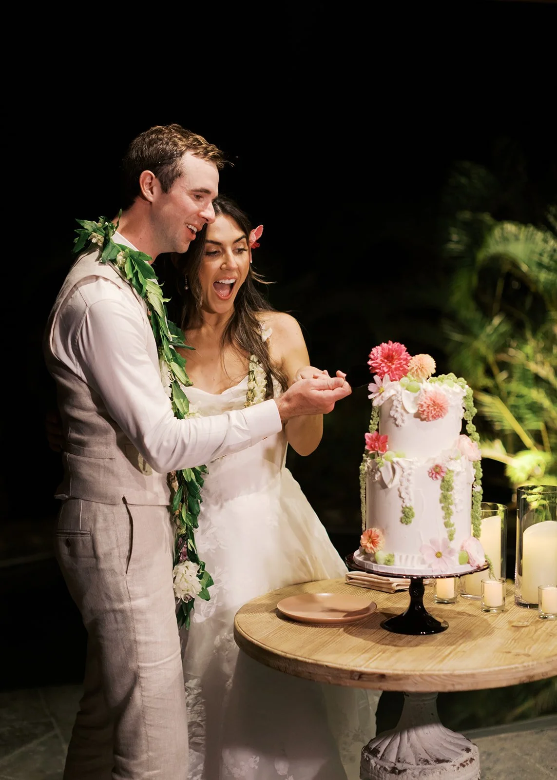 A bride and groom cut a wedding cake together at their wedding reception, standing by a table with candles and flowers, with a dark background