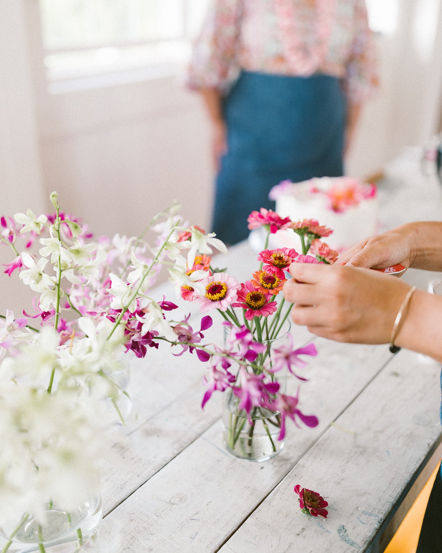 Several small flower arrangements with pink, white, and purple flowers in glass vases on a white table. In the background, a person wearing a floral top and a blue apron is preparing flowers.