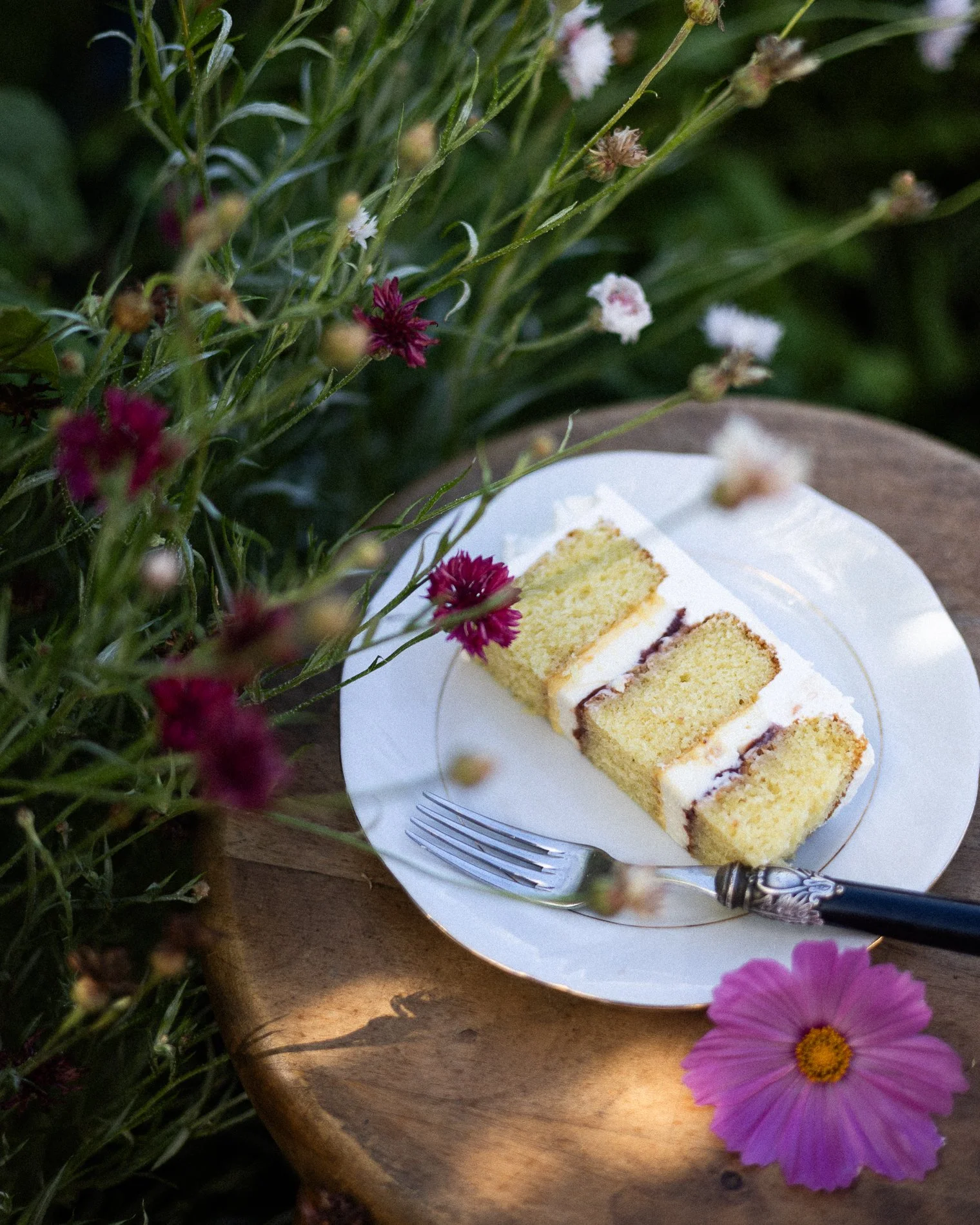 Slices of vanilla cake with cream filling and chocolate, on a white plate, with a fork and knife, surrounded by colorful flowers, on a wooden surface.