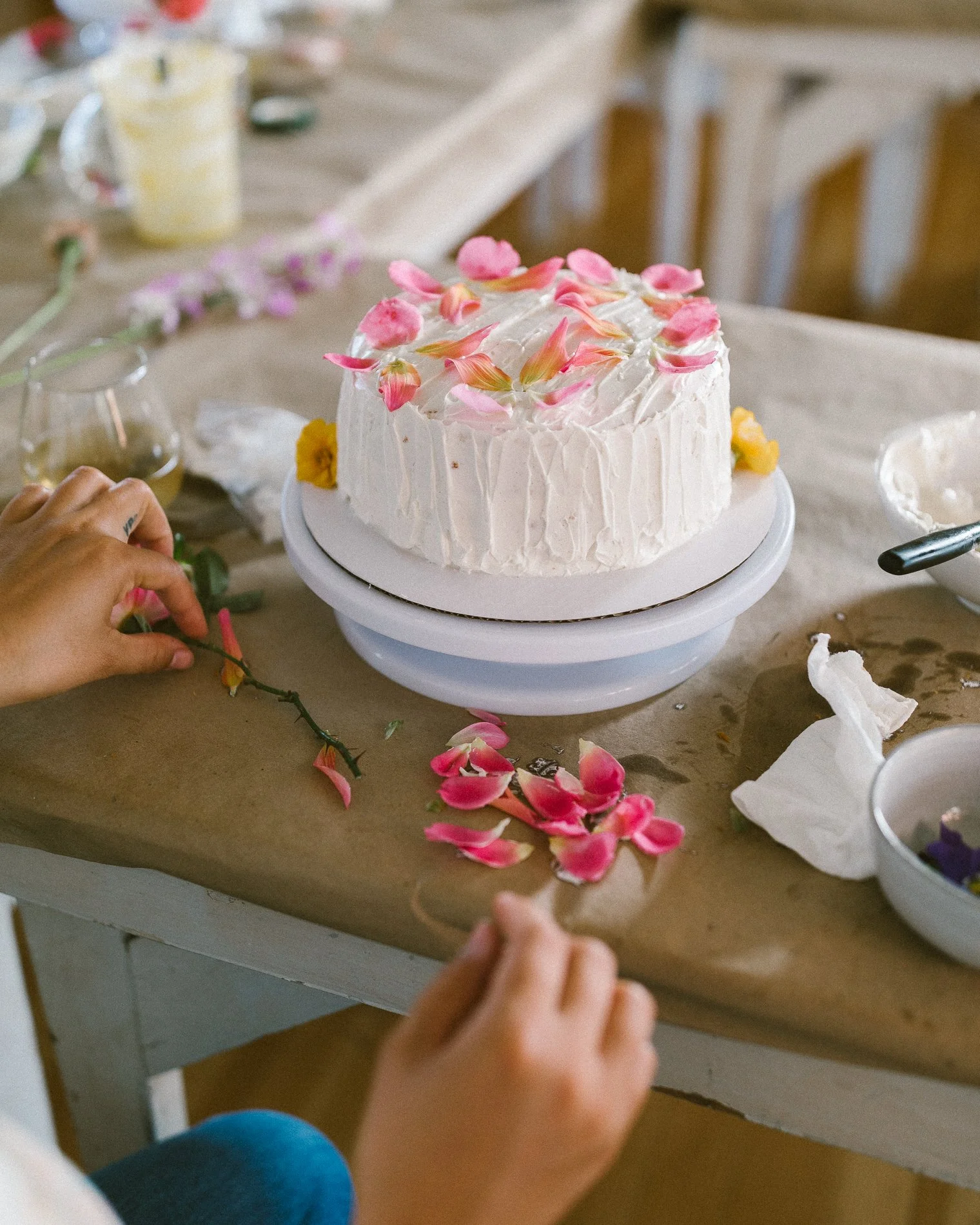 A birthday cake decorated with pink and yellow flower petals on a white icing frosting, sitting on a cake stand on a cluttered table with scattered flower petals, paper towels, and other baking supplies.