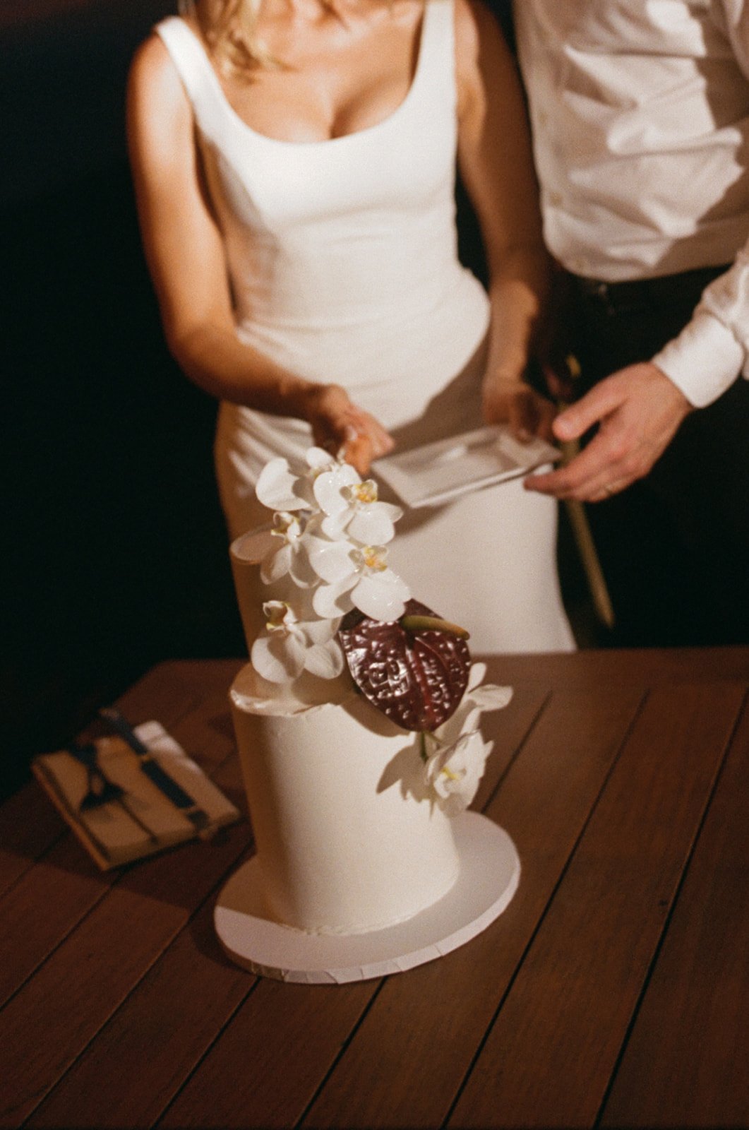 A woman in a white dress and a man in a white shirt are cutting a wedding cake decorated with orchids and a chocolate heart on top.