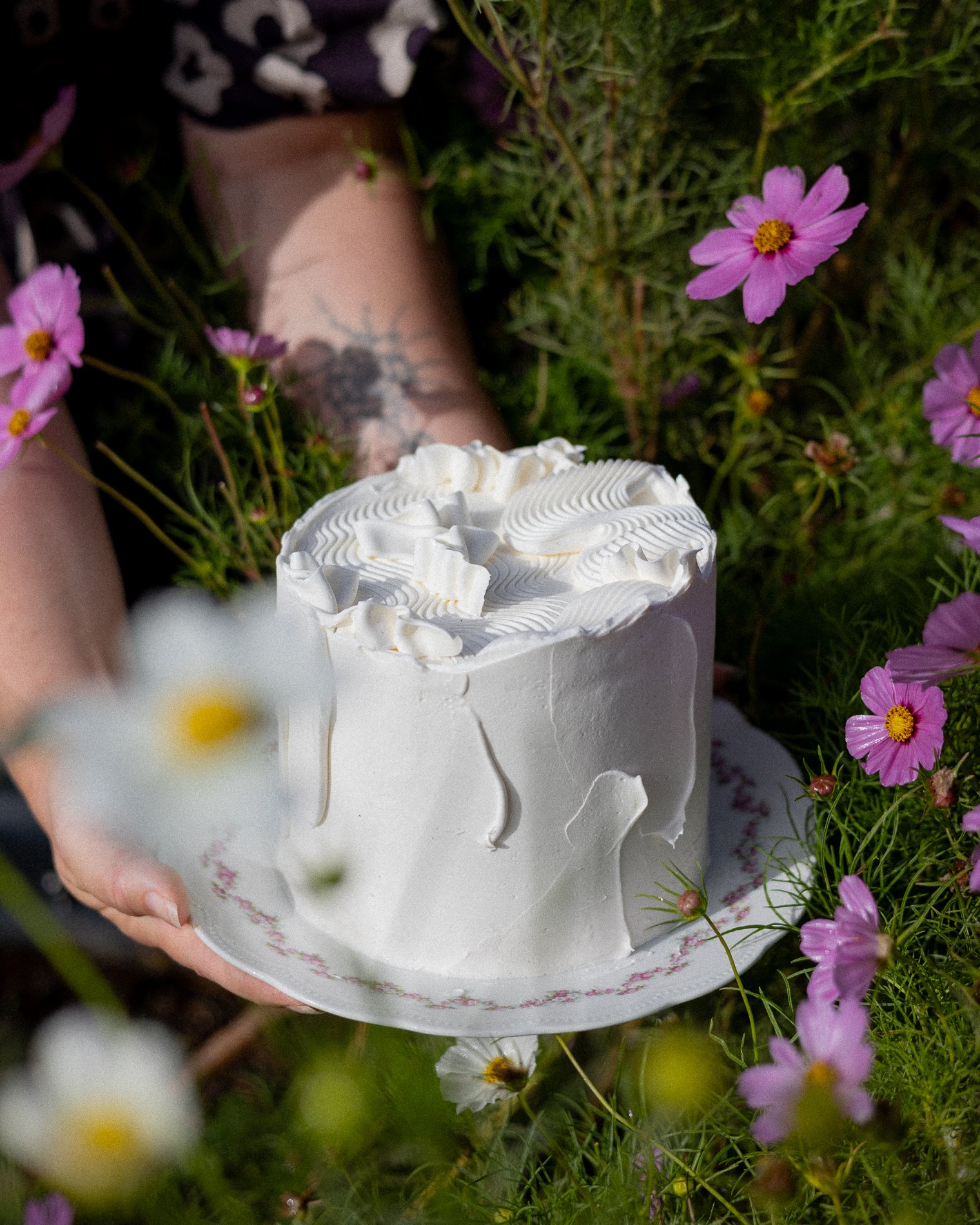 A person's hand holding a white cake with decorative white icing inside a ring of purple flowers with yellow centers, set outdoors on green grass.