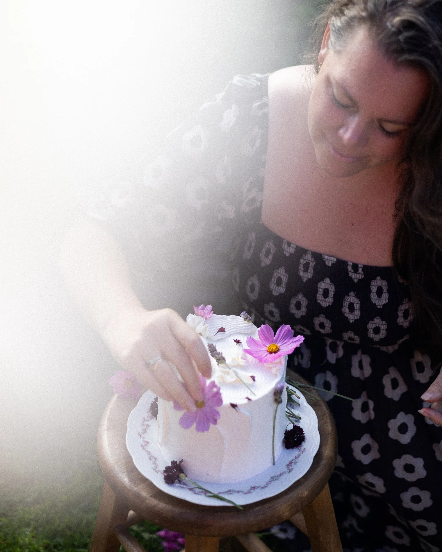 A woman with long dark hair, wearing a black and white floral dress, is decorating a white cake with pink and purple flowers on a wooden table outdoors.
