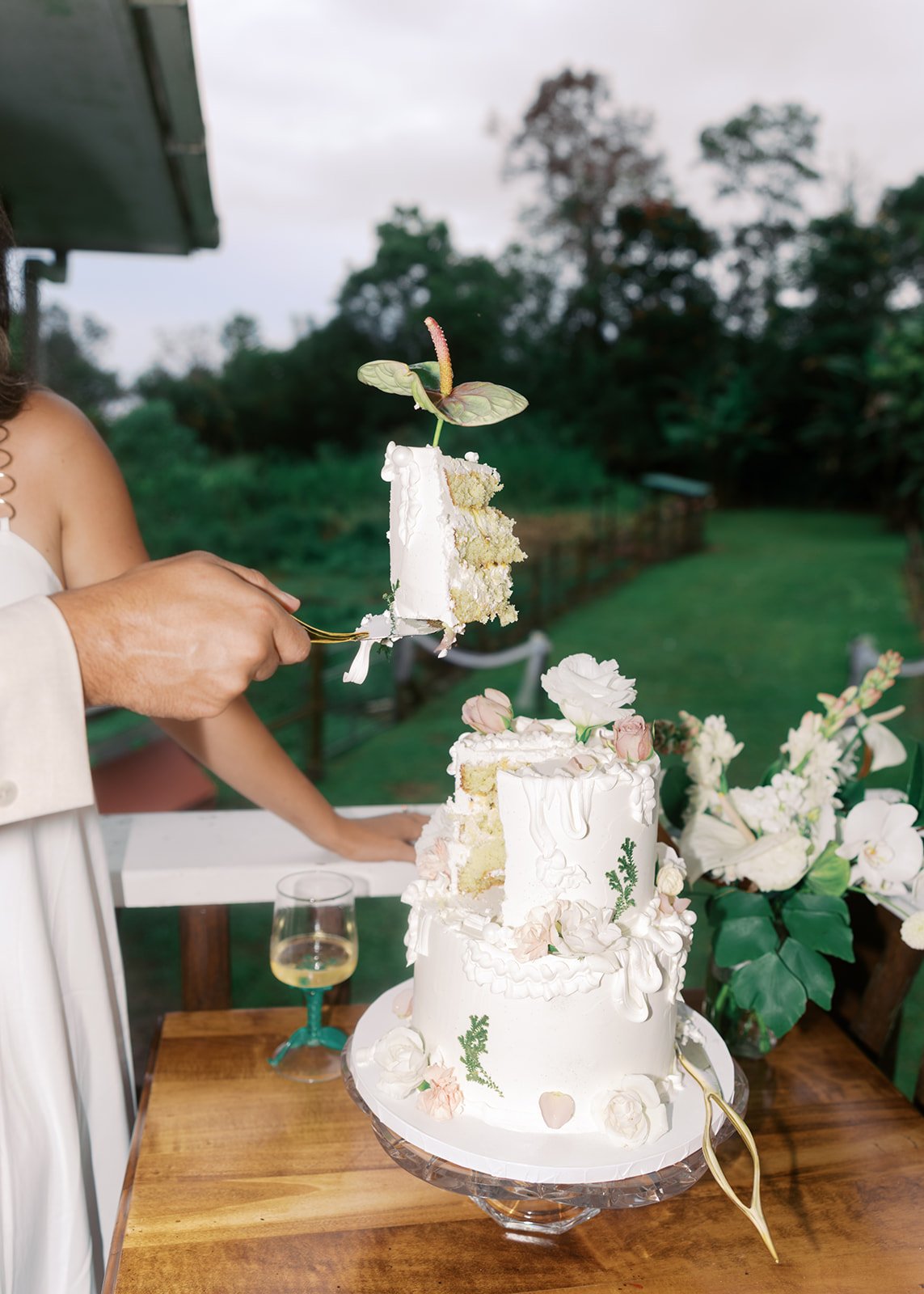 A person is cutting a three-tiered wedding cake decorated with white and pink flowers and greenery, outdoors with trees and a cloudy sky in the background. There are flowers and a glass of champagne on the table.