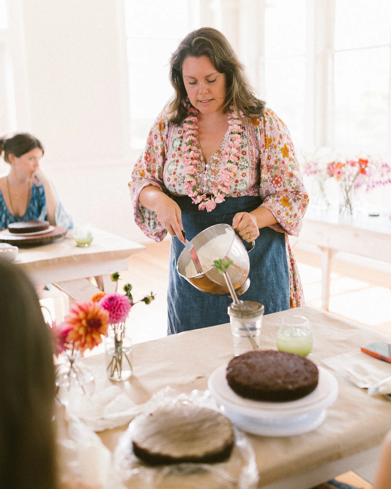 A woman with brown hair wearing a floral shirt and pink flower lei pouring a liquid into a glass on a table set for a celebration, with another woman in the background and a chocolate cake in the foreground.