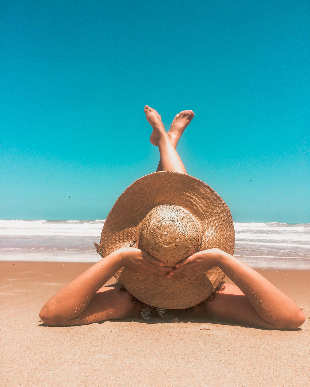 woman lying on beach with straw hat on with legs in the air at the beach