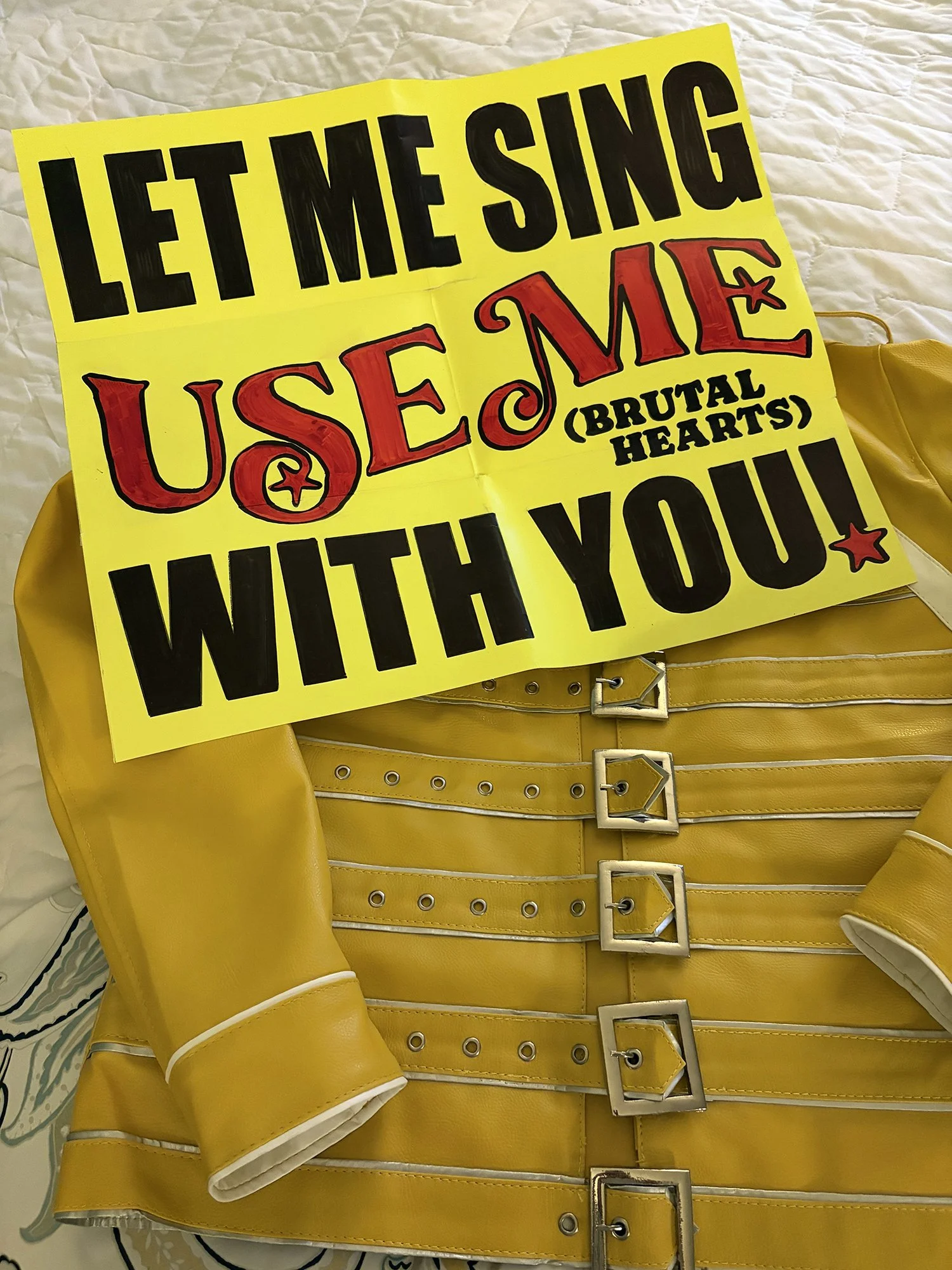 Freddie Mercury yellow buckled jacket with a neon poster board laying over it that says "Let me sing Use Me with you!"