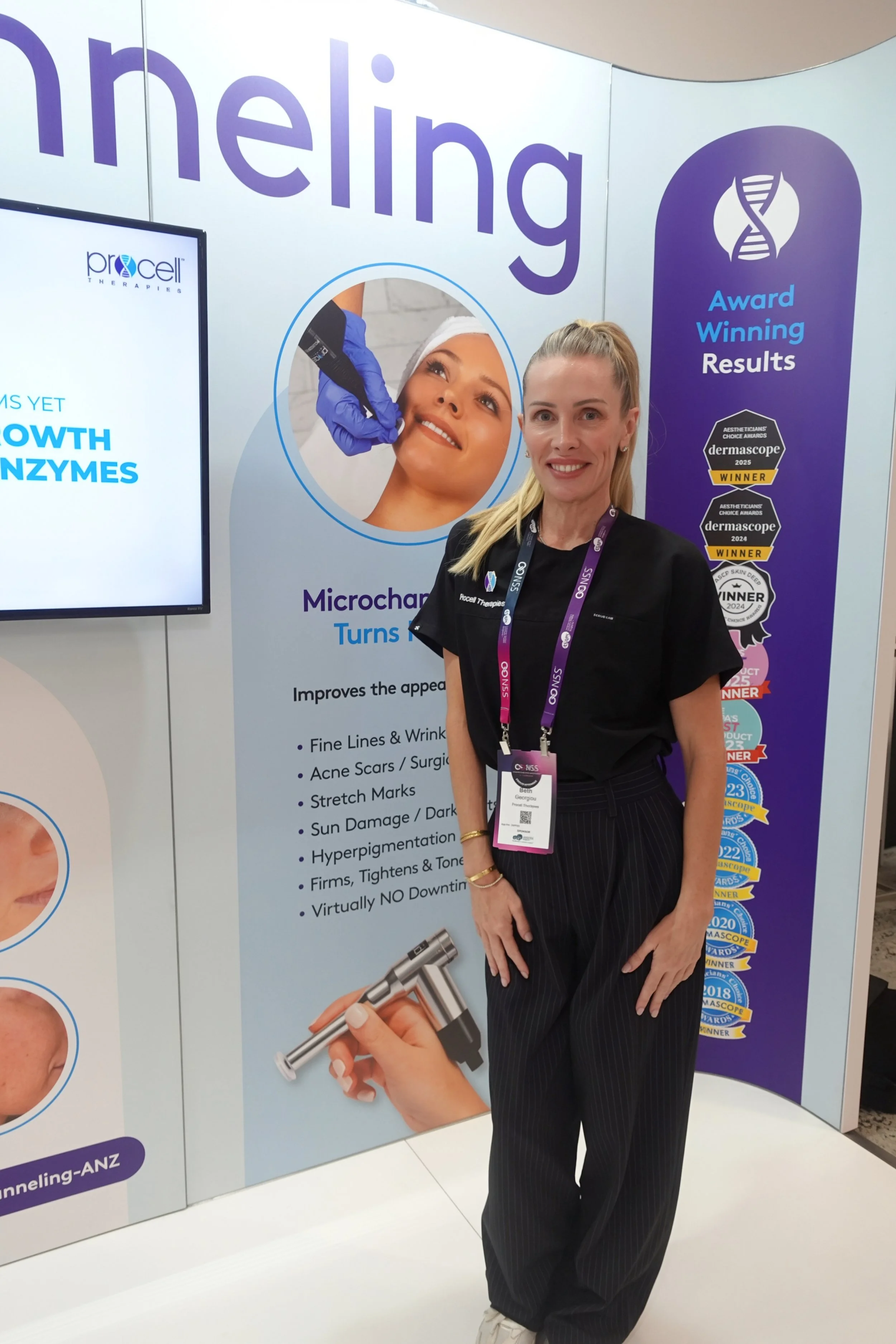 A woman standing in front of a display booth at a conference, wearing a black shirt and pants with a lanyard badge. The booth has promotional posters about skincare treatments, awards, and results, along with images of a woman receiving a facial treatment and a hand holding a microneedling device.
