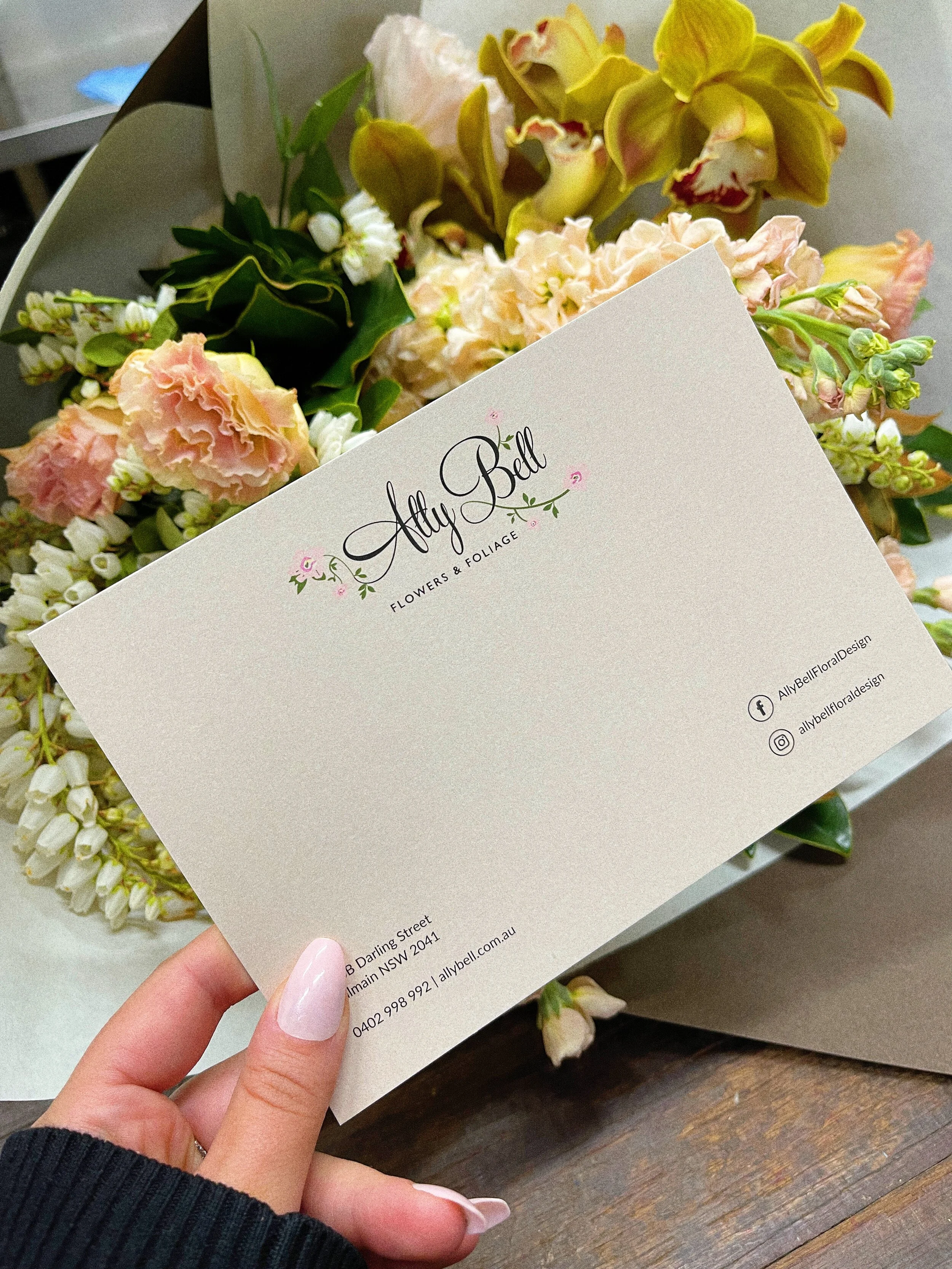 A hand holding a business card for Ally Bell Flowers & Foliage with a background of assorted flowers including pink blossoms, white small flowers, and yellow flowers.