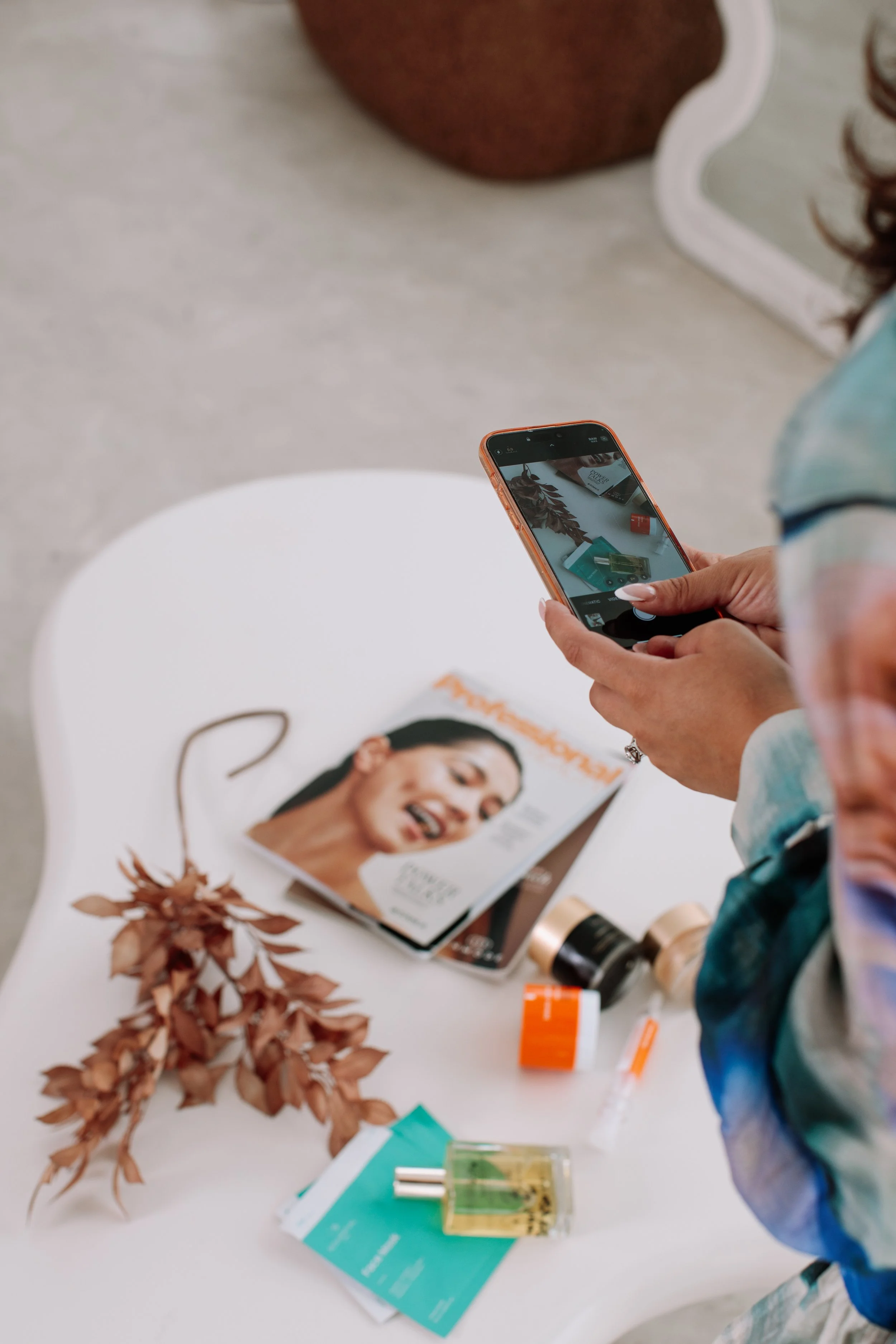 A person taking a photo of a magazine on a white table using a smartphone, with various skincare products and a dried plant nearby.