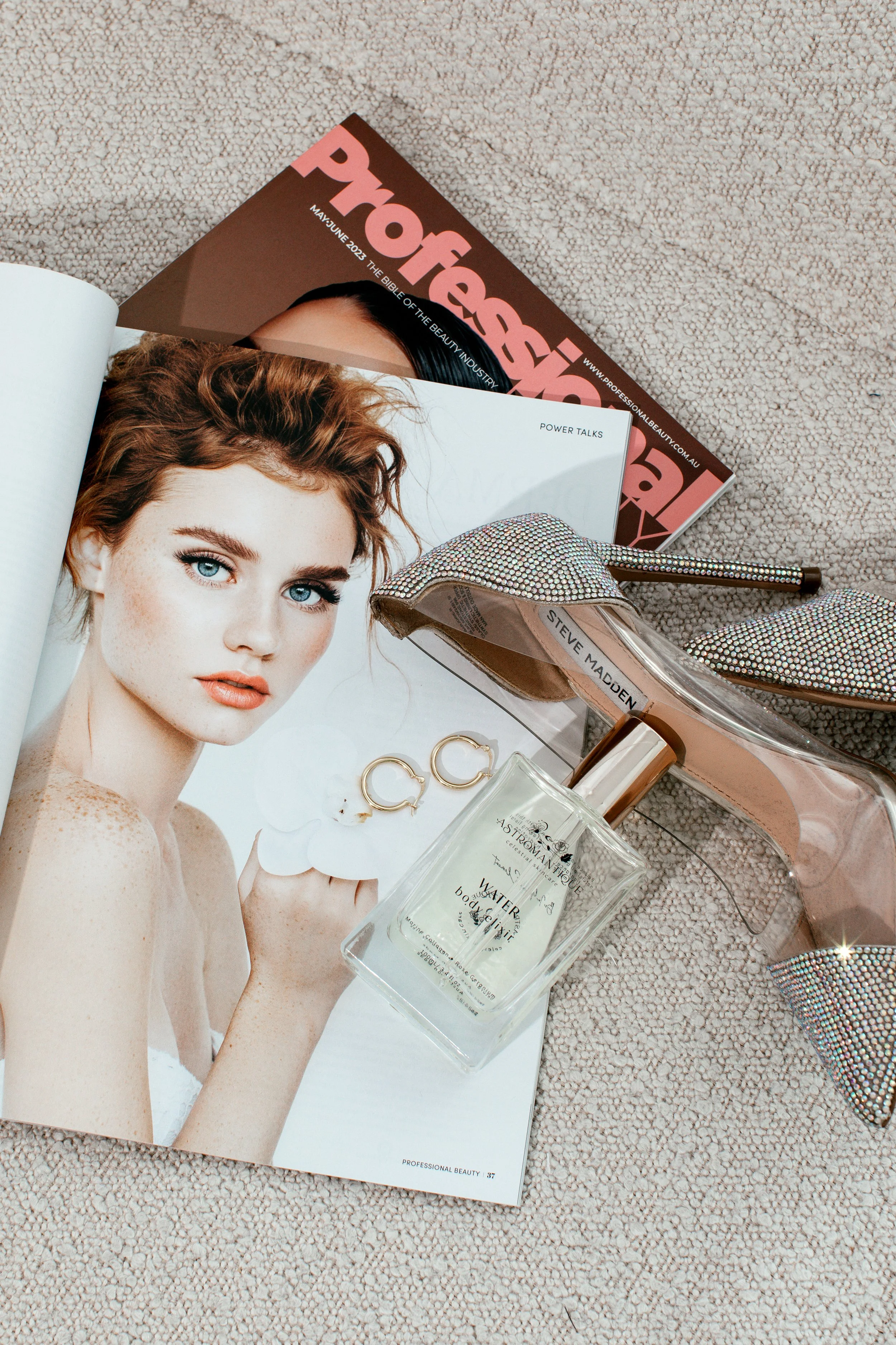 Beauty magazine, open magazine with a woman’s face on one page, perfume bottle, sparkly high heel shoes, and jewelry on a beige textured surface.