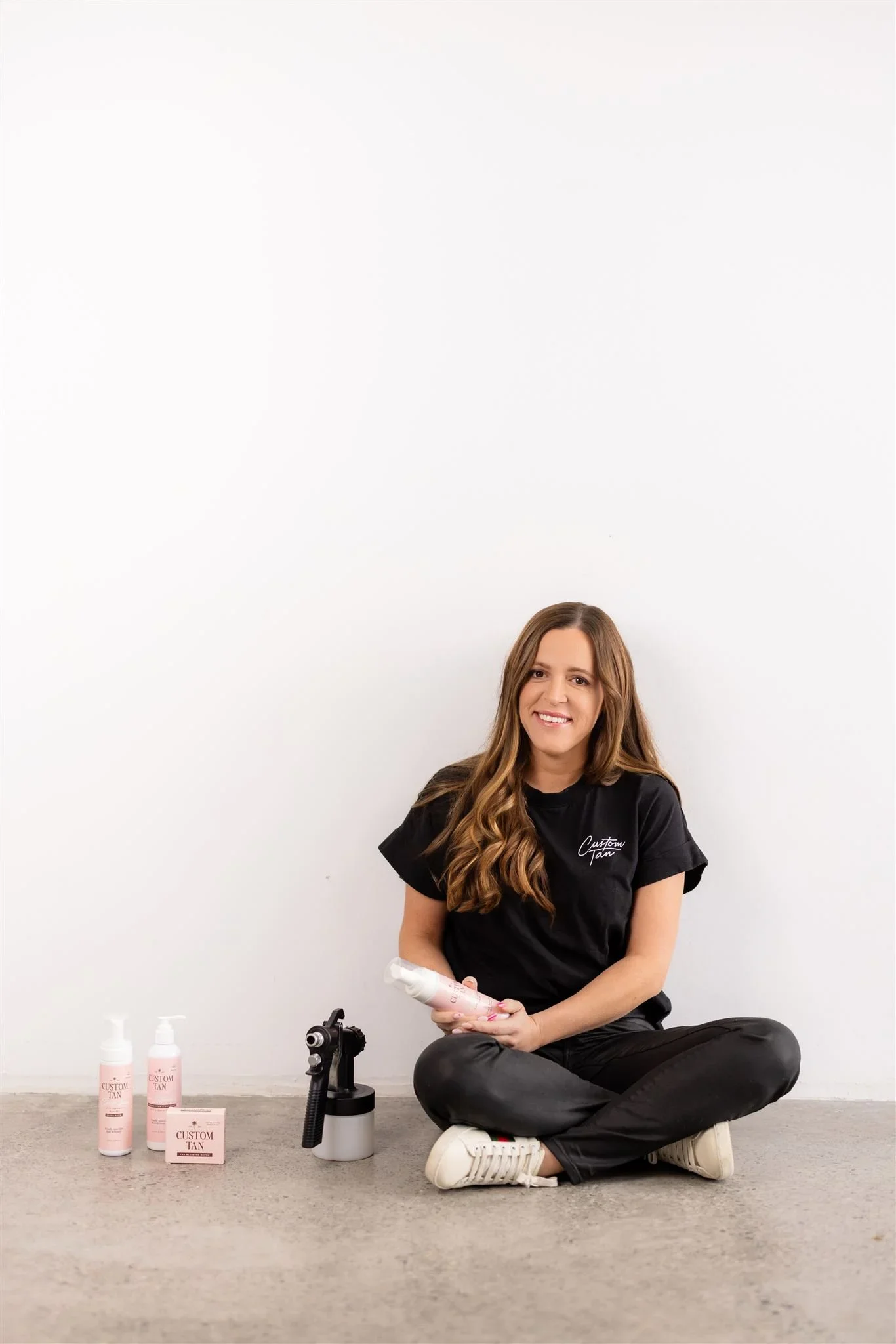 A woman sitting on the floor with a tattoo gun in front of her, holding a tattoo ink bottle, surrounded by tattoo supplies including bottles labeled 'Custom Tan,' and a small pink tray with tattoo ink. She is wearing a black t-shirt and black pants, sitting against a plain white wall.
