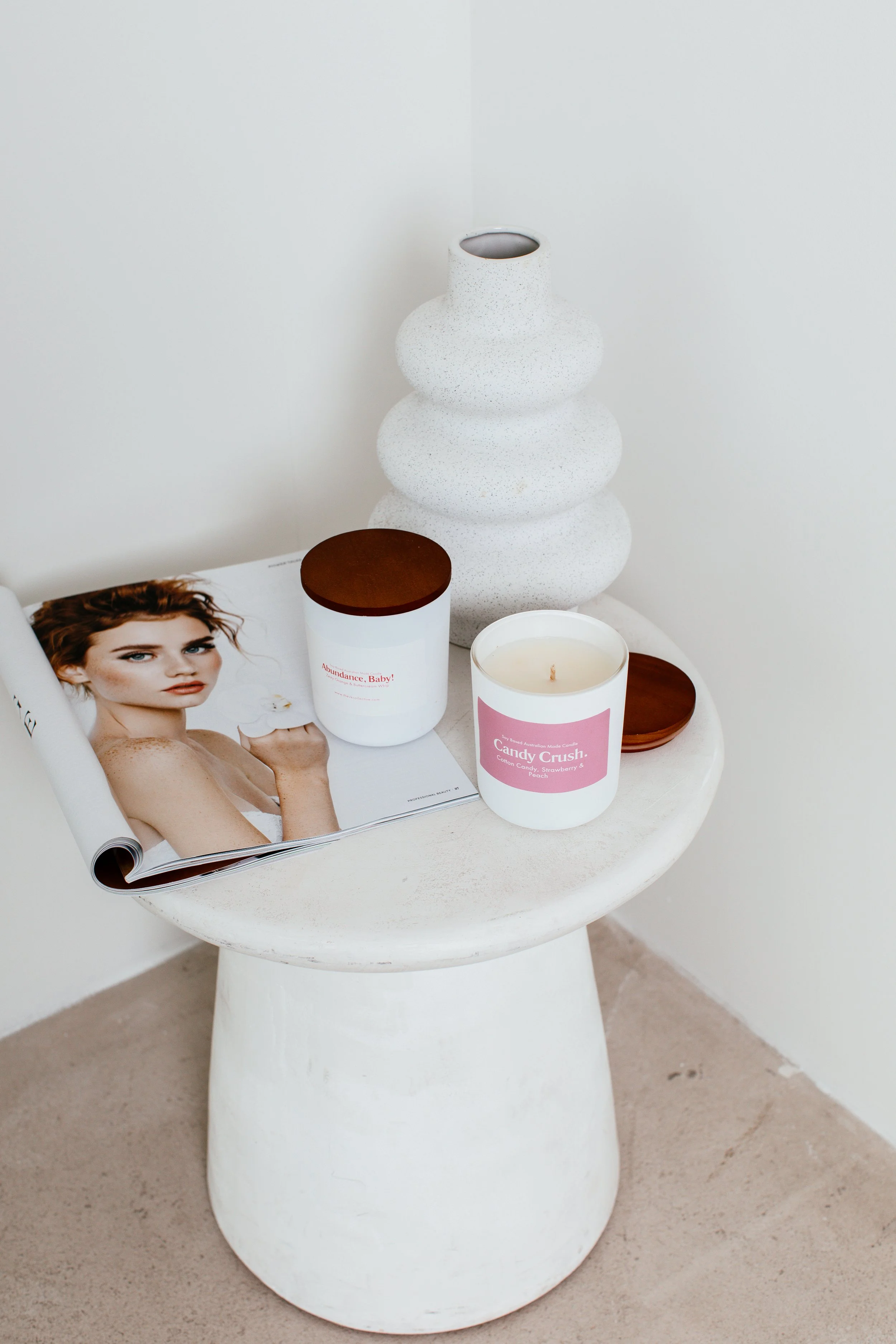 A white round table with a large white textured vase, a candle labeled 'Candy Crush,' and a magazine with a woman on the cover, set against a plain white wall.