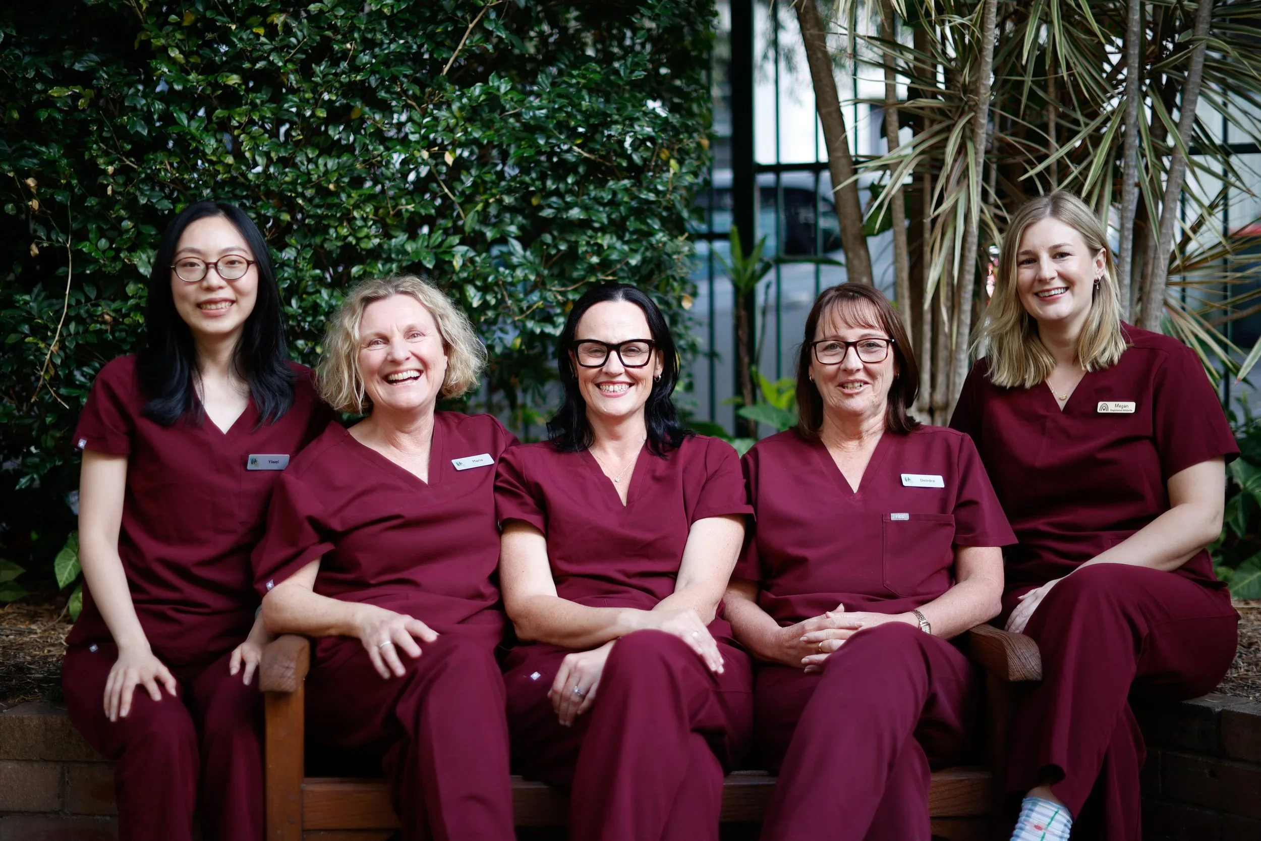 Group of five women in maroon uniforms sitting outdoors in front of a dense green leafy background, smiling at the camera.