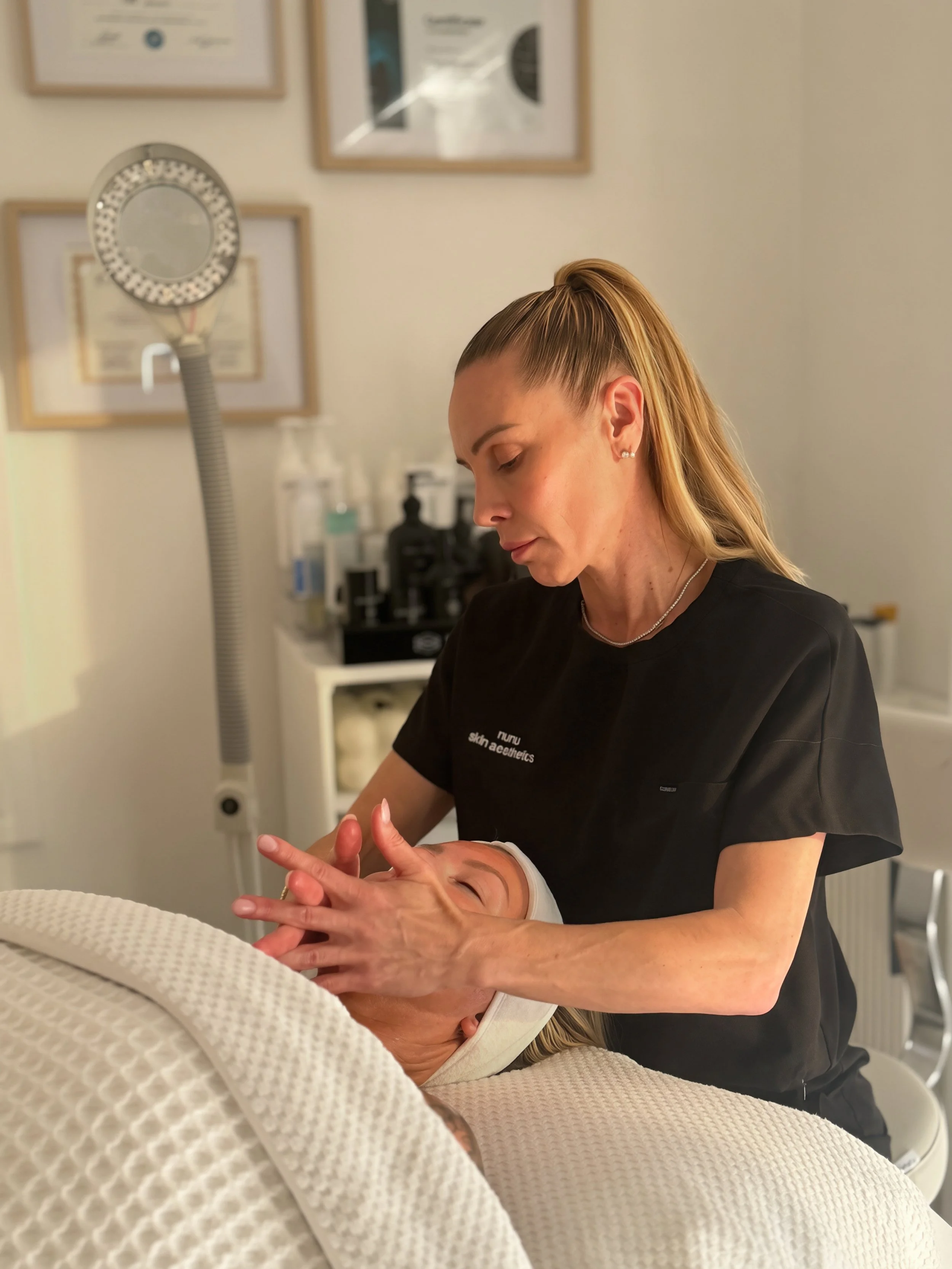 A female healthcare worker giving a facial or skincare treatment to a woman lying on a treatment bed in a clinic or spa setting.
