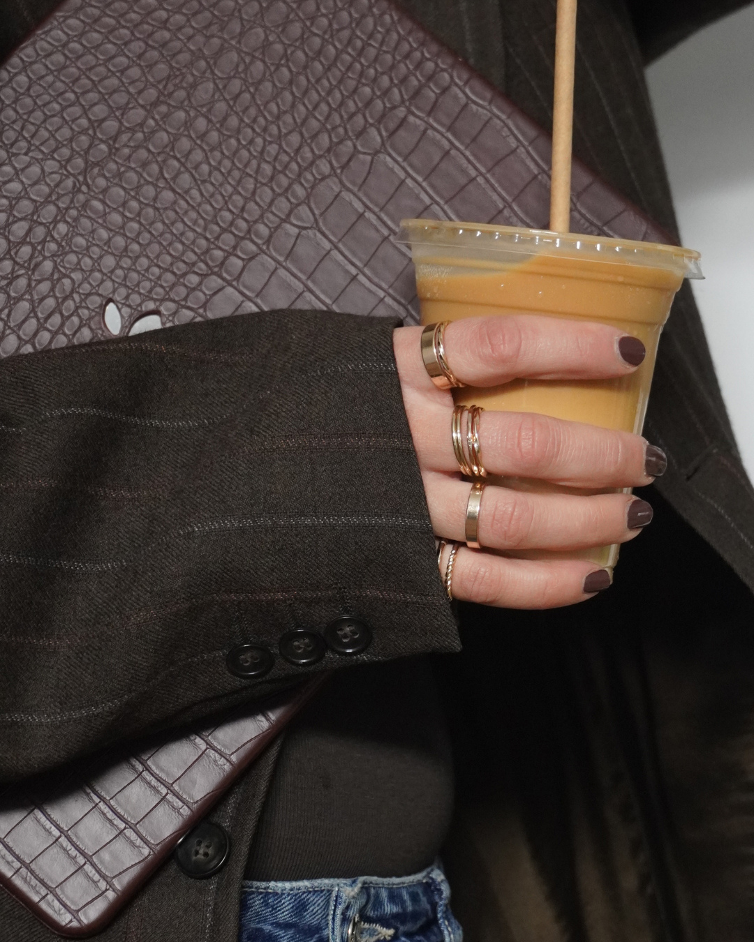 Person holding a plastic cup of iced coffee with a straw, wearing multiple rings and dark-colored nail polish, dressed in a dark blazer and blue jeans, with a brown textured purse or wallet partially visible.