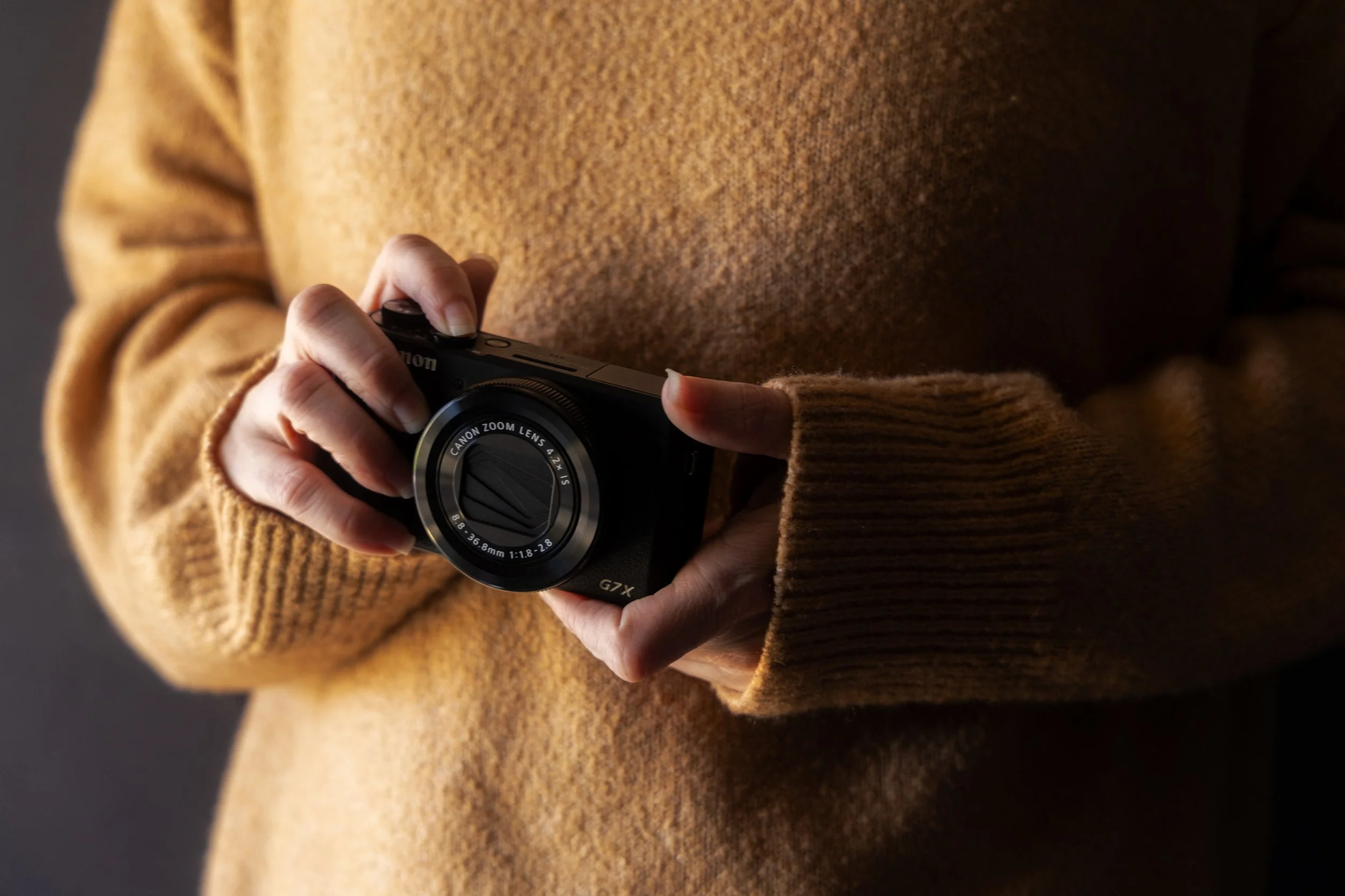 Person wearing a brown sweater holding a Canon point-and-shoot camera.