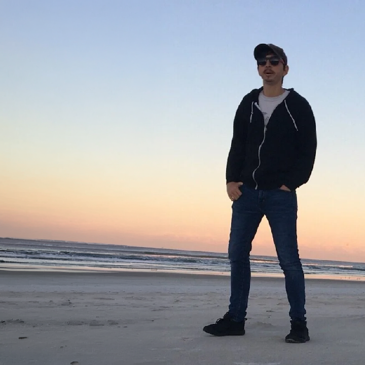 D. Max(@D_Max_HS) standing on a beach at sunset, wearing a black jacket, jeans, and sunglasses.