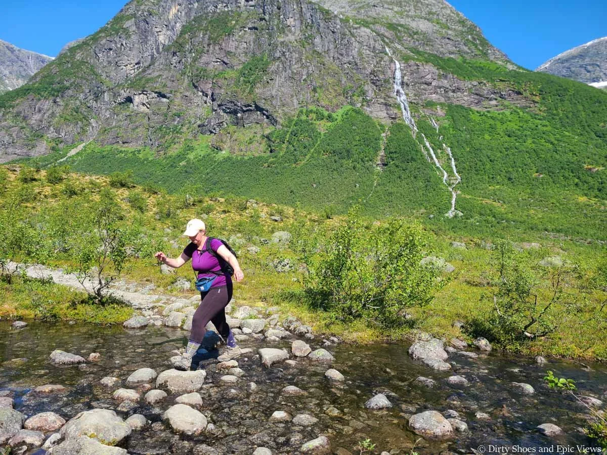 A hiker rock hops across a stream in front of waterfalls and mountain views on the Austerdalsbreen hike in Norway