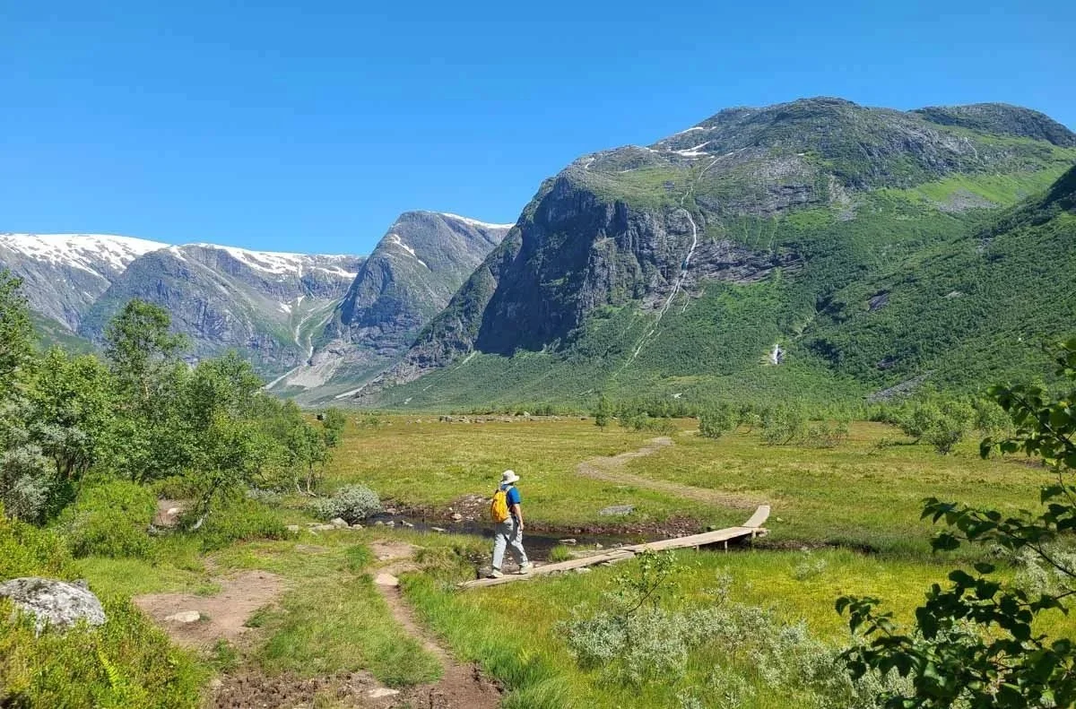 A hiker crosses a small footbridge in a grassy valley between mountains on the Austerdalsbreen hike in Norway