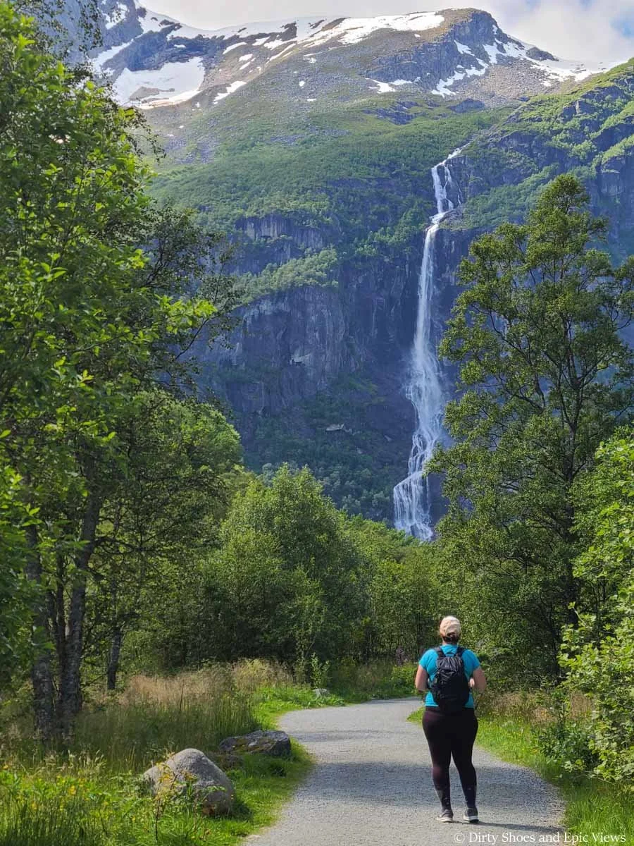 A hiker walks along a flat paved path towards a view of a waterfall on the Briksdal Glacier trail in Norway