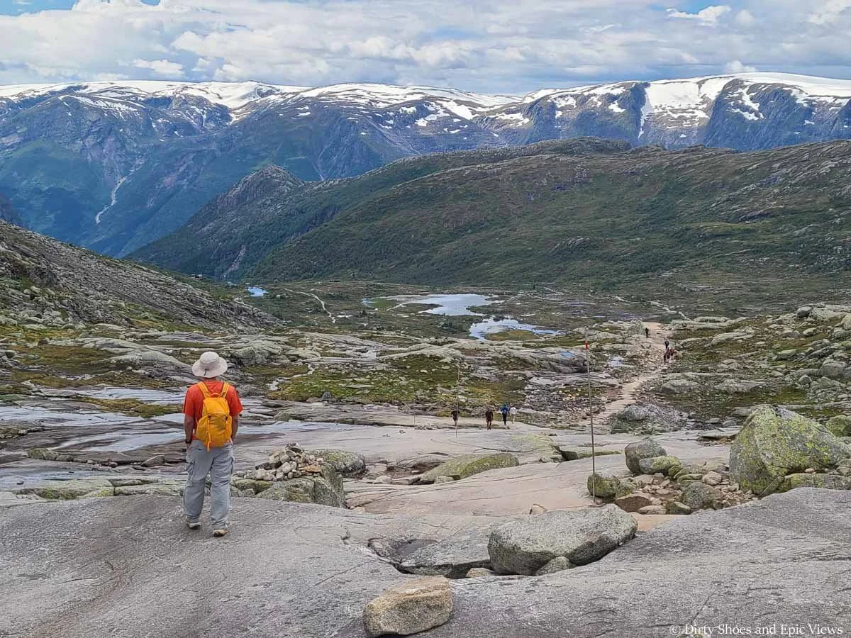 A hiker descends down a rocky slab towards views of a large meadow with lakes and snow capped mountains on the Trolltunga trail