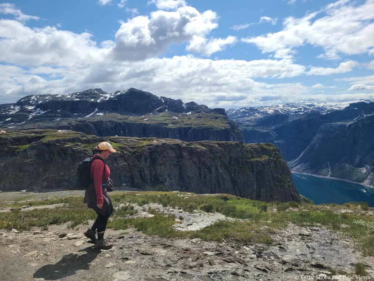 A hiker walks in front of views of steep cliffs and a blue lake on the Trolltunga trail
