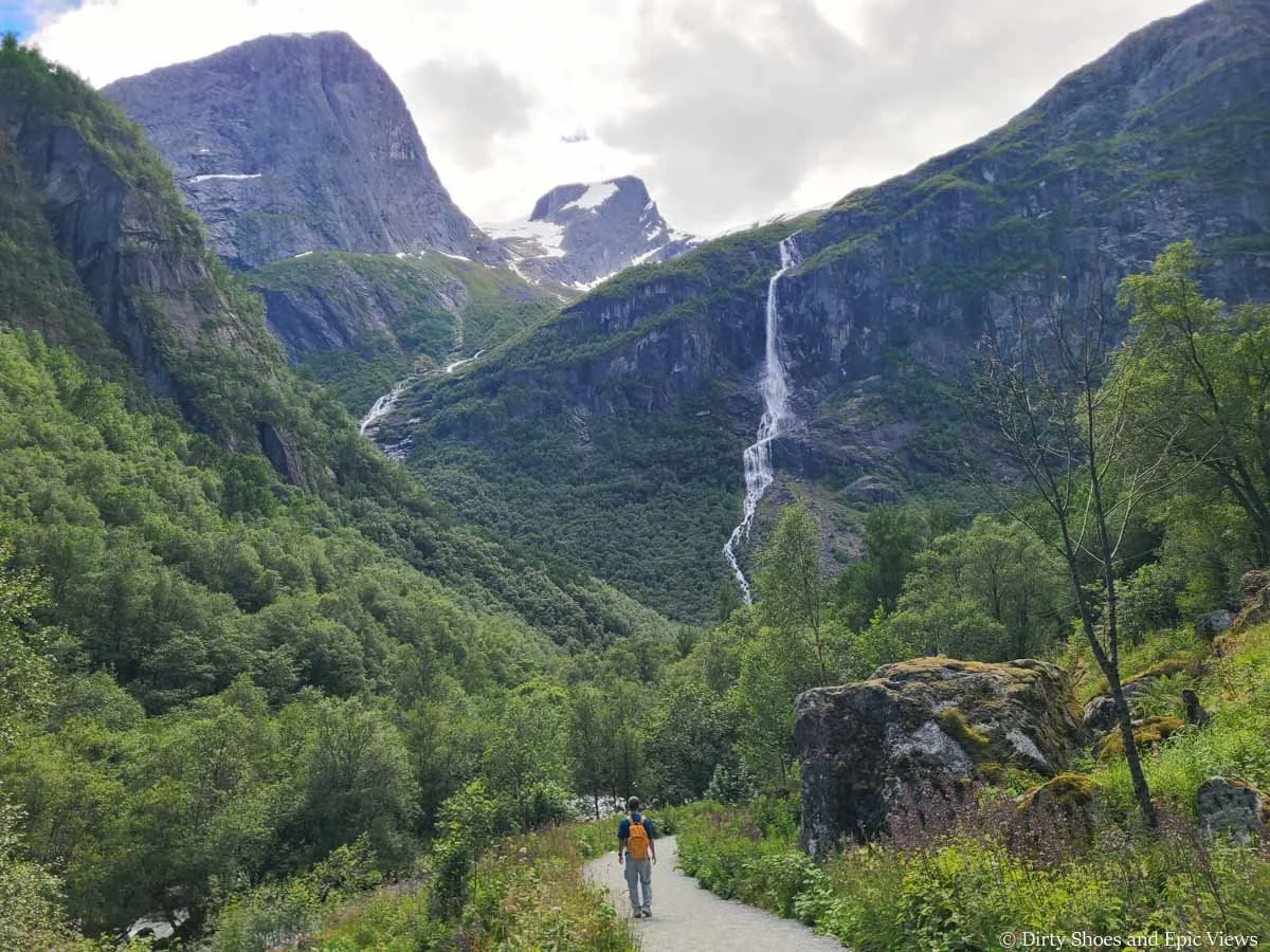A hiker walks a flat path towards views of waterfalls cascading down mountain faces on the Briksdal Glacier trail in Norway