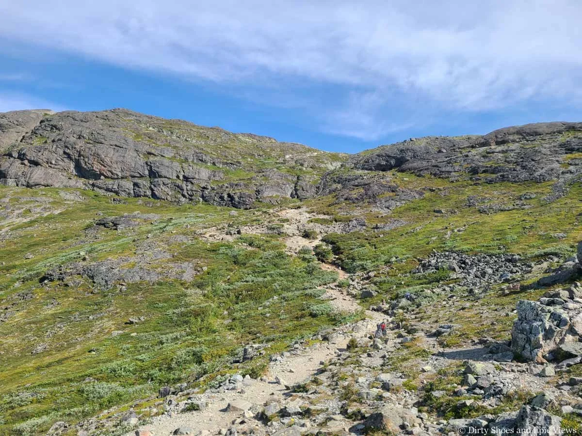 A narrow dirt trail weaves through a grassy meadow towards a rock wall on the Besseggen Ridge trail in Norway