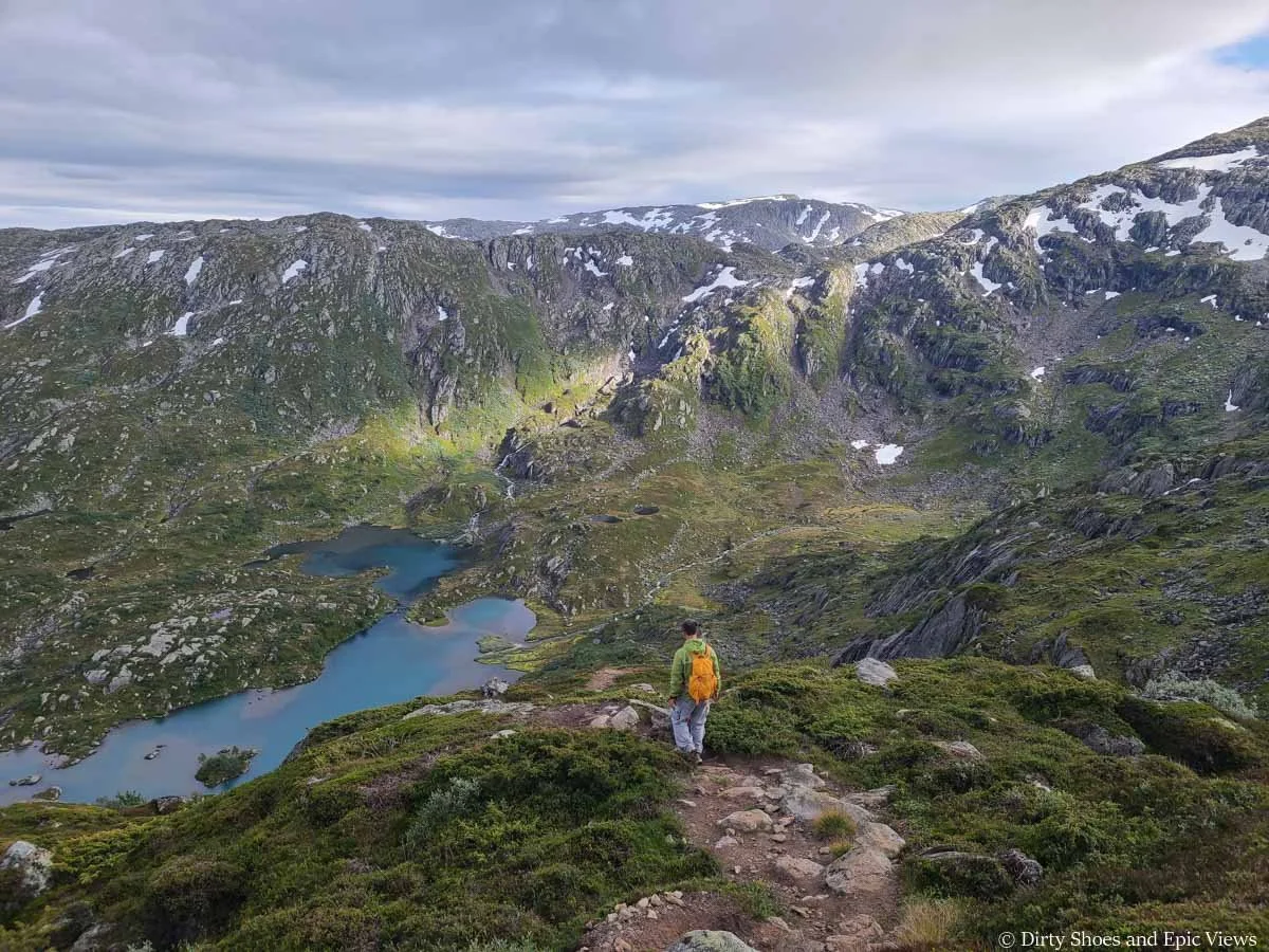 A hiker walks down a rocky path towards views of a basin with blue lakes and waterfalls on the Reinanuten trail in Norway