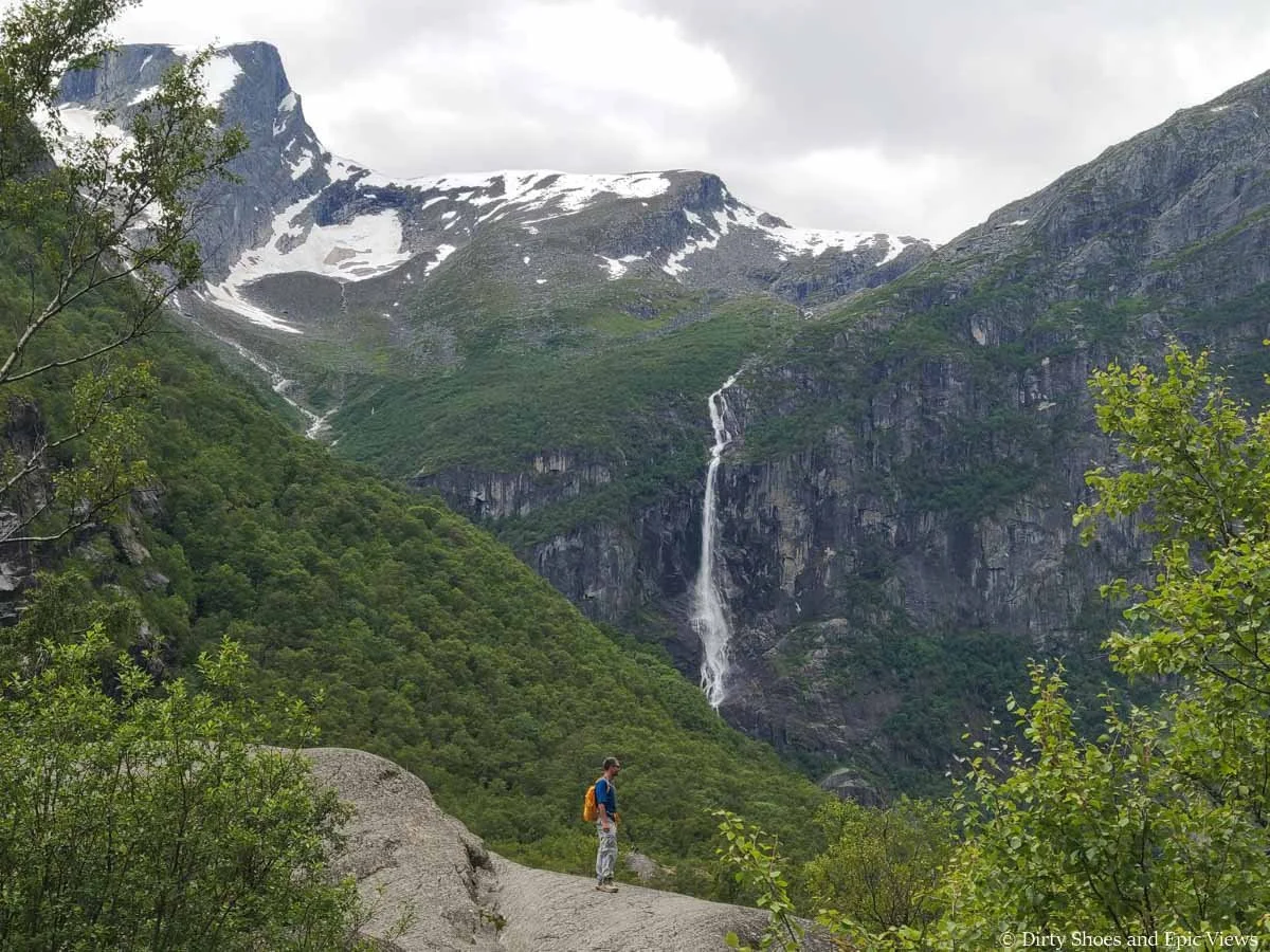 A hiker stands on a boulder overlooking mountains and a waterfall on the Briksdalsbreen hike in Norway
