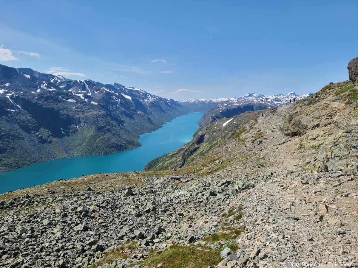 A narrow path runs along a cliff edge over a blue lake and mountain views on Besseggen Ridge in Norway