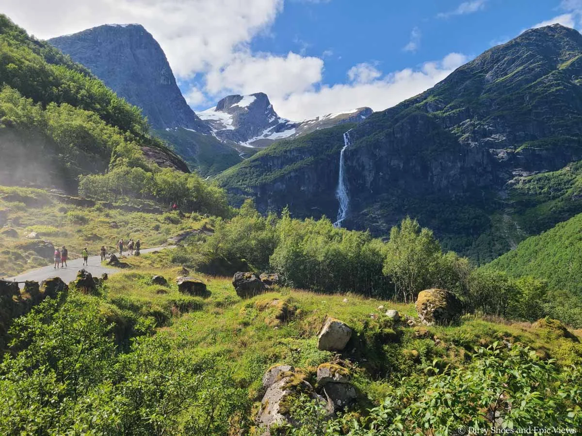 A crowded hiking path crosses a meadow towards a waterfall on the Briksdal Glacier trail in Norway