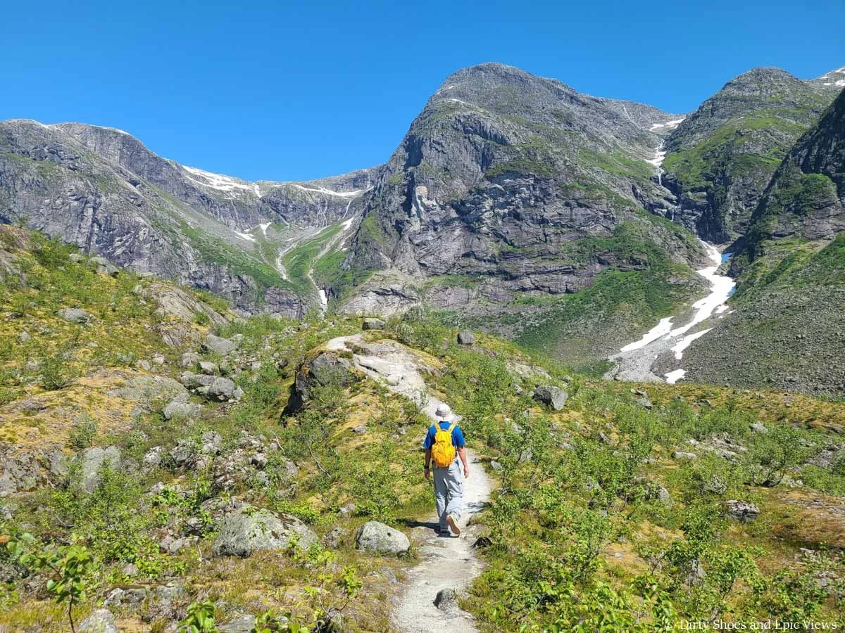 A hiker walks along a narrow dirt path towards the top of a hill with mountain views on the Austerdalsbreen hike