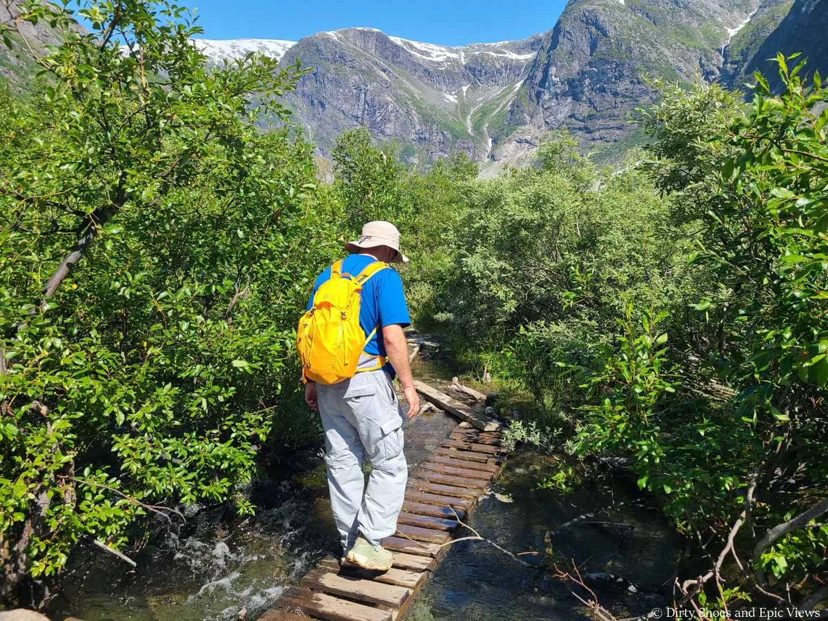A hiker crosses a footbridge through a stream and brush on the Austerdalsbreen hike in Norway