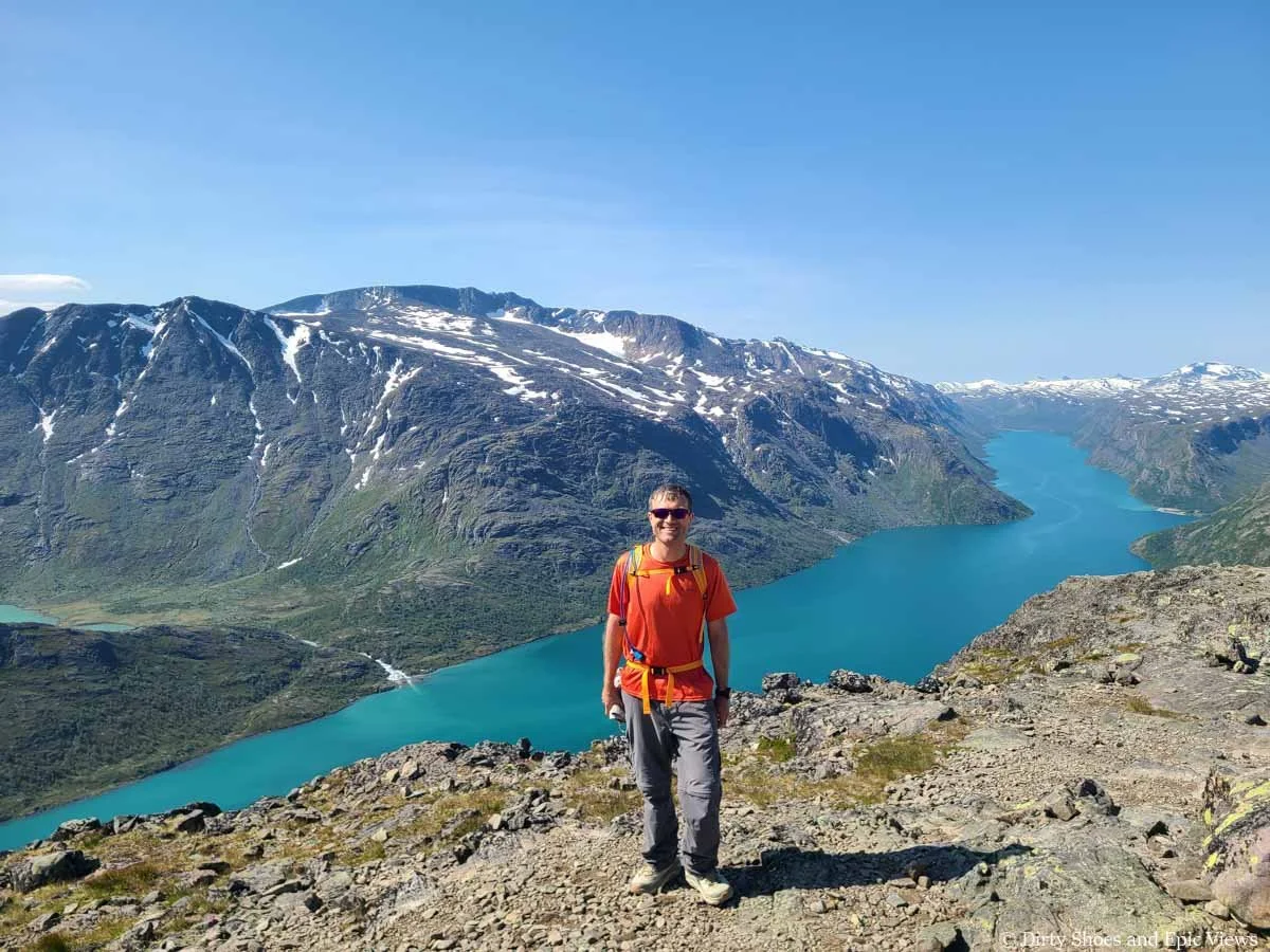 A hiker smiles at the camera in front of views of mountains and a turquoise lake below from the Besseggen Ridge hike in Norway