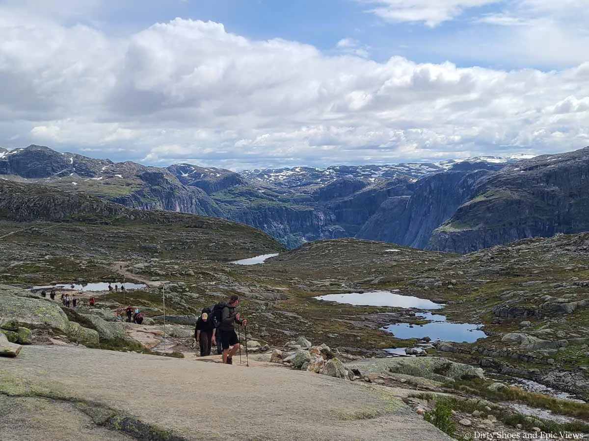 A line of hikers ascend a trail with mountain and lake views on the Trolltunga hike