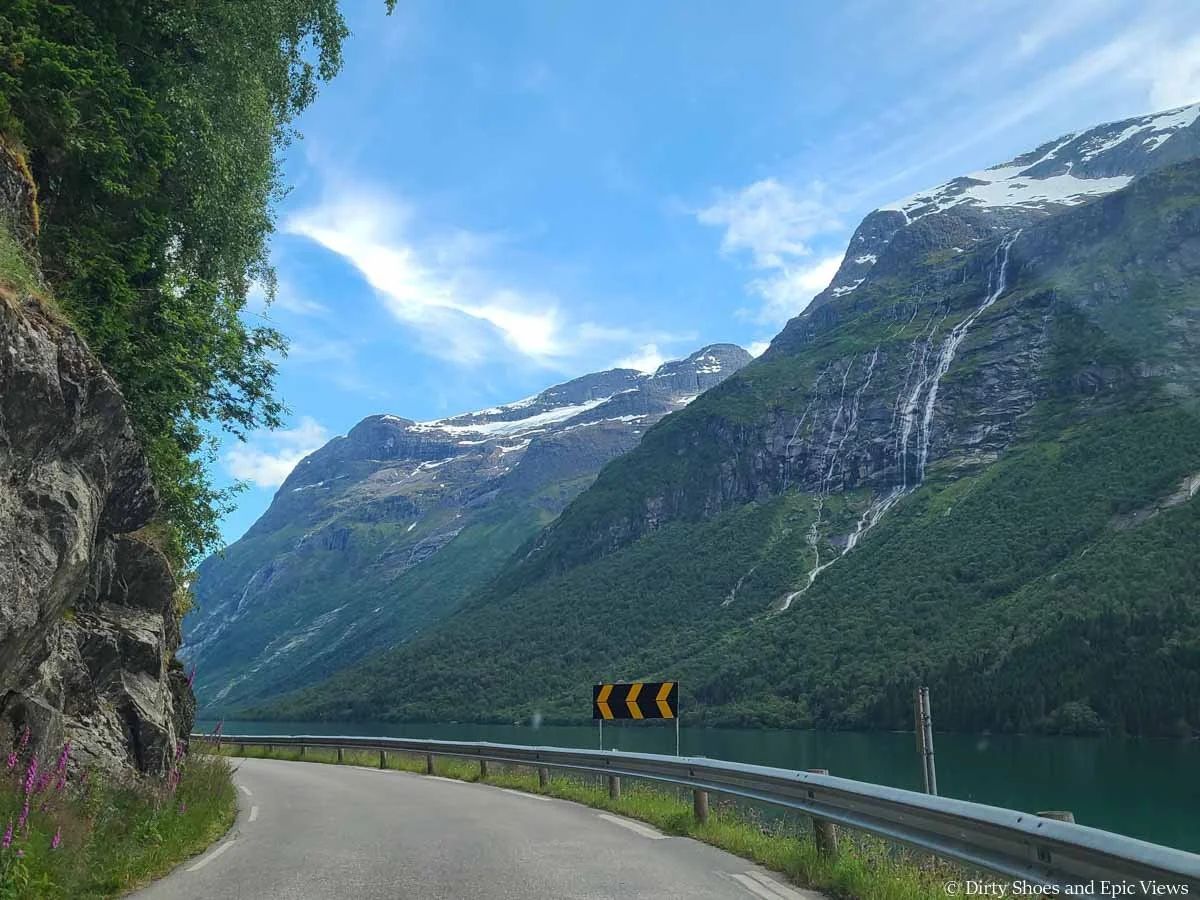 A road makes a blind turn around a rock wall with mountain views on the road to Lovatnet in Norway