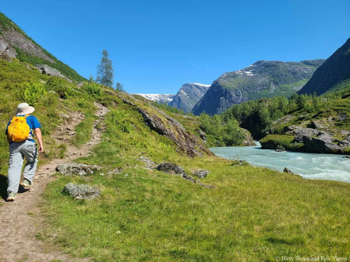A hiker ascends a narrow dirt path by a river on the Austerdalsbreen hike in Norway
