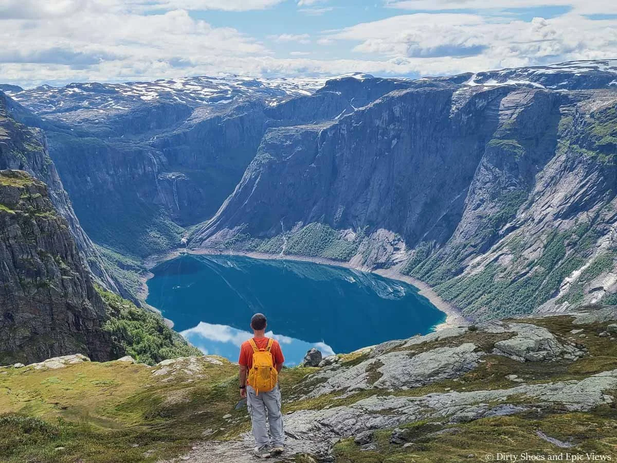 A hiker stands overlooking views of a blue lake surrounded by steep rocky cliffs and waterfalls on the Trolltunga hike