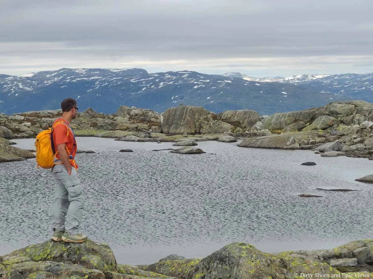 A hiker stands in front of a lake backdropped by a mountain ridge at the Reinanuten viewpoint in Norway